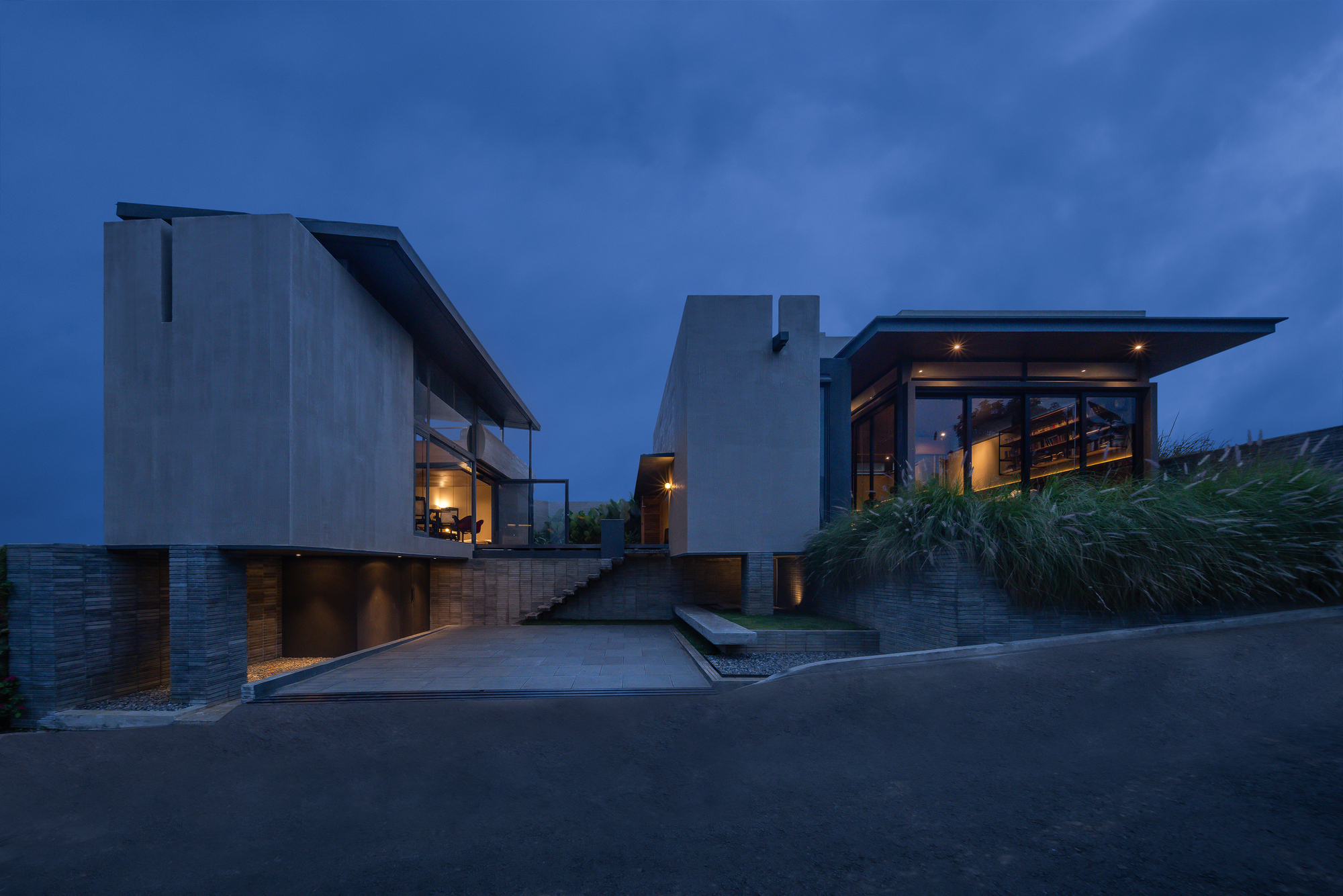 Street view of stacked concrete volumes with timber base and planted grasses at dusk