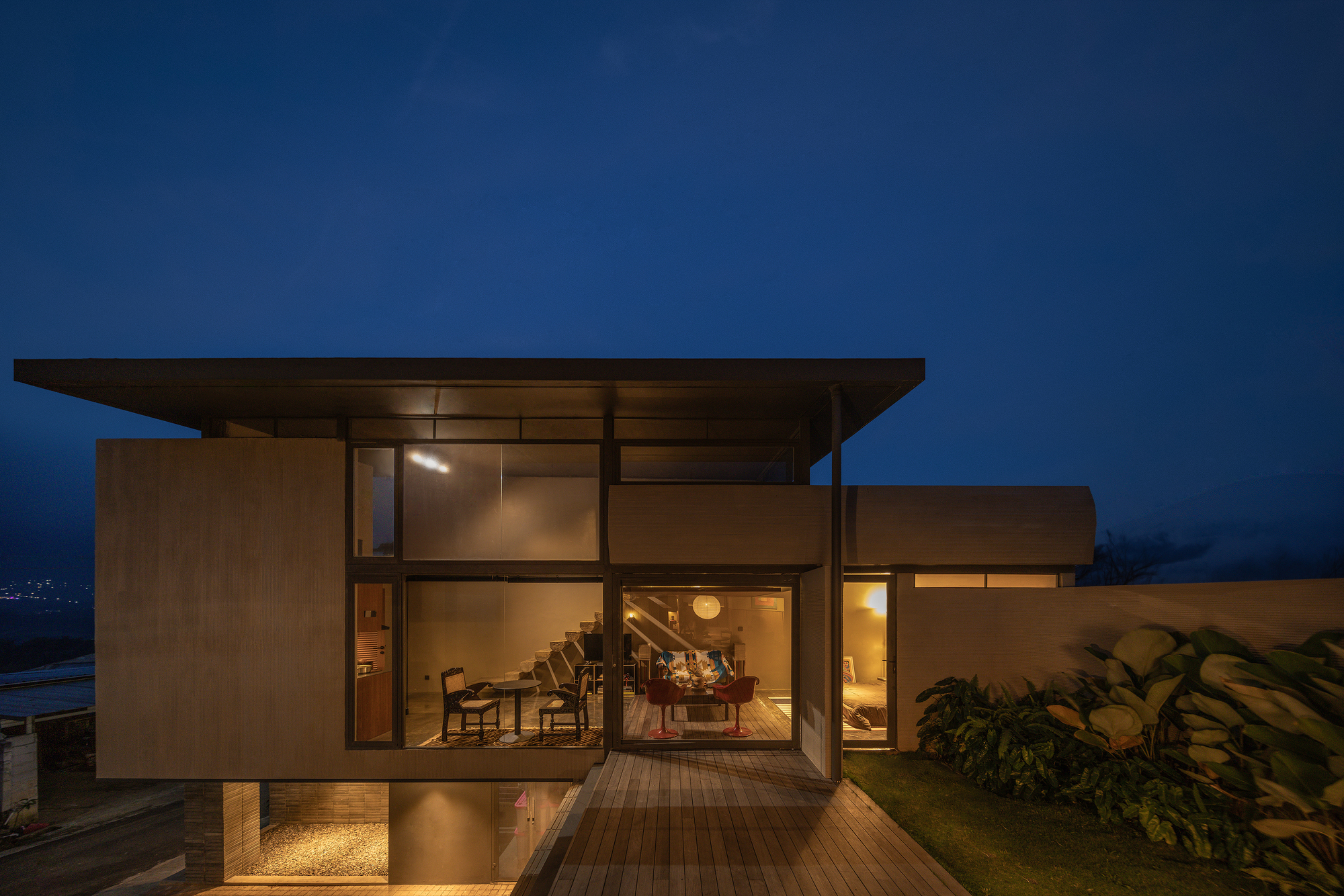 Two-level residence with cantilevered flat roof and timber deck extending over a planted garden at dusk