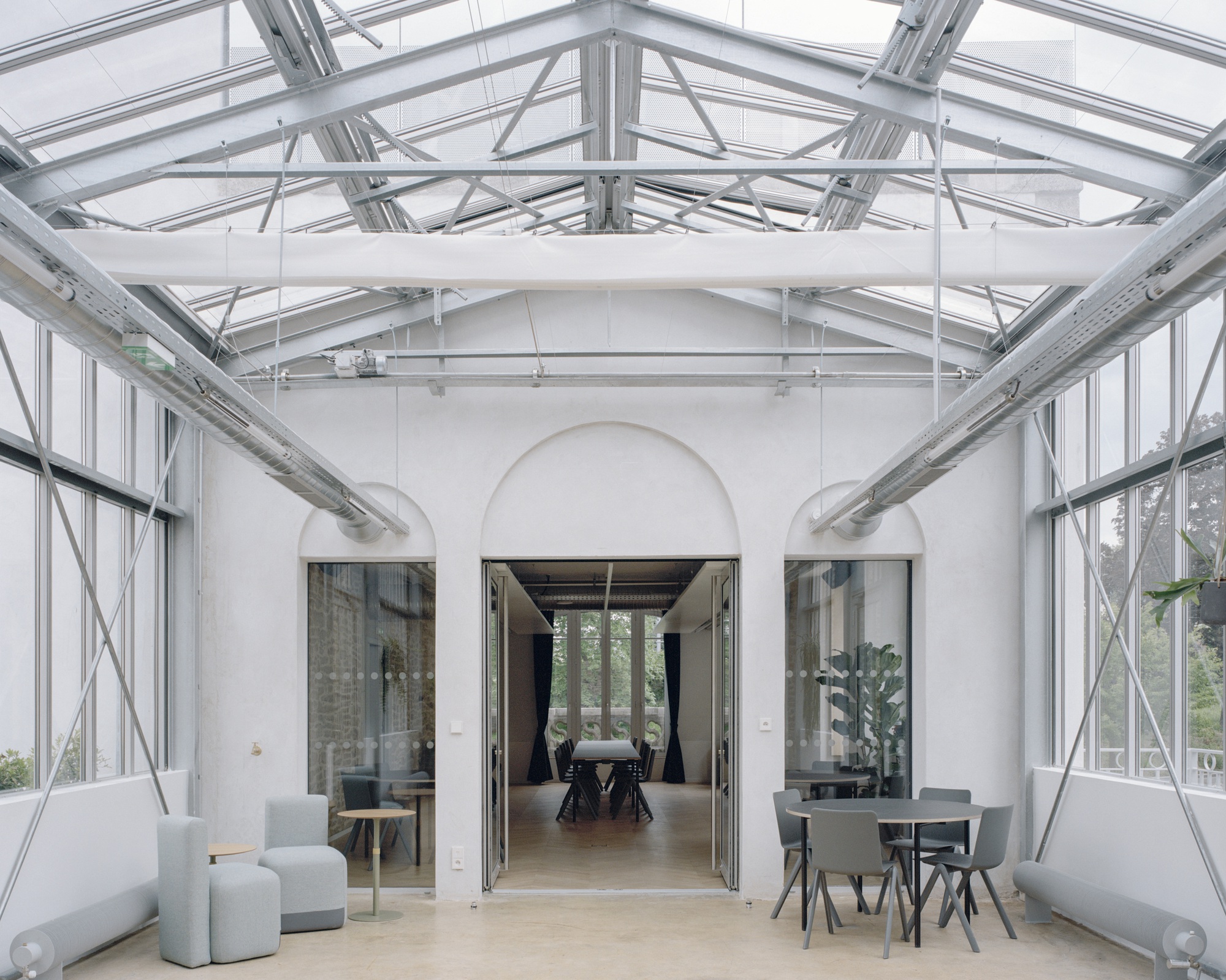 Glazed conservatory space with steel truss roof, arched opening and scattered seating under natural light