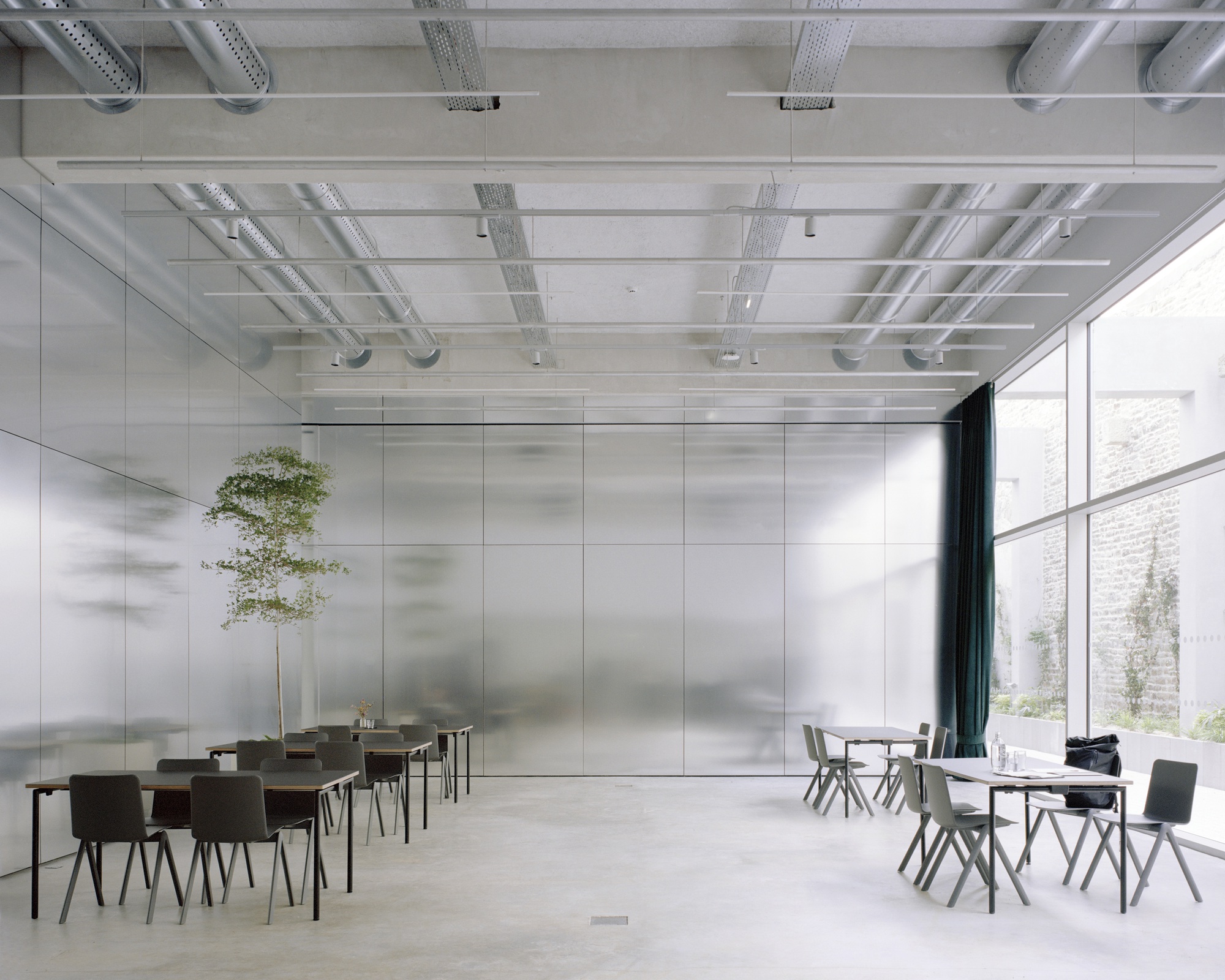 Interior hall with polished stainless steel wall panels, exposed ceiling ducts, and scattered dining tables near a tall glazed wall