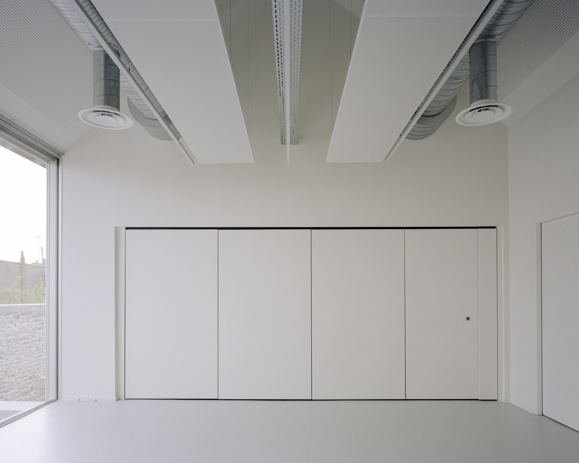 Open room with exposed ceiling ducts and perforated acoustic panels above flush storage cabinets