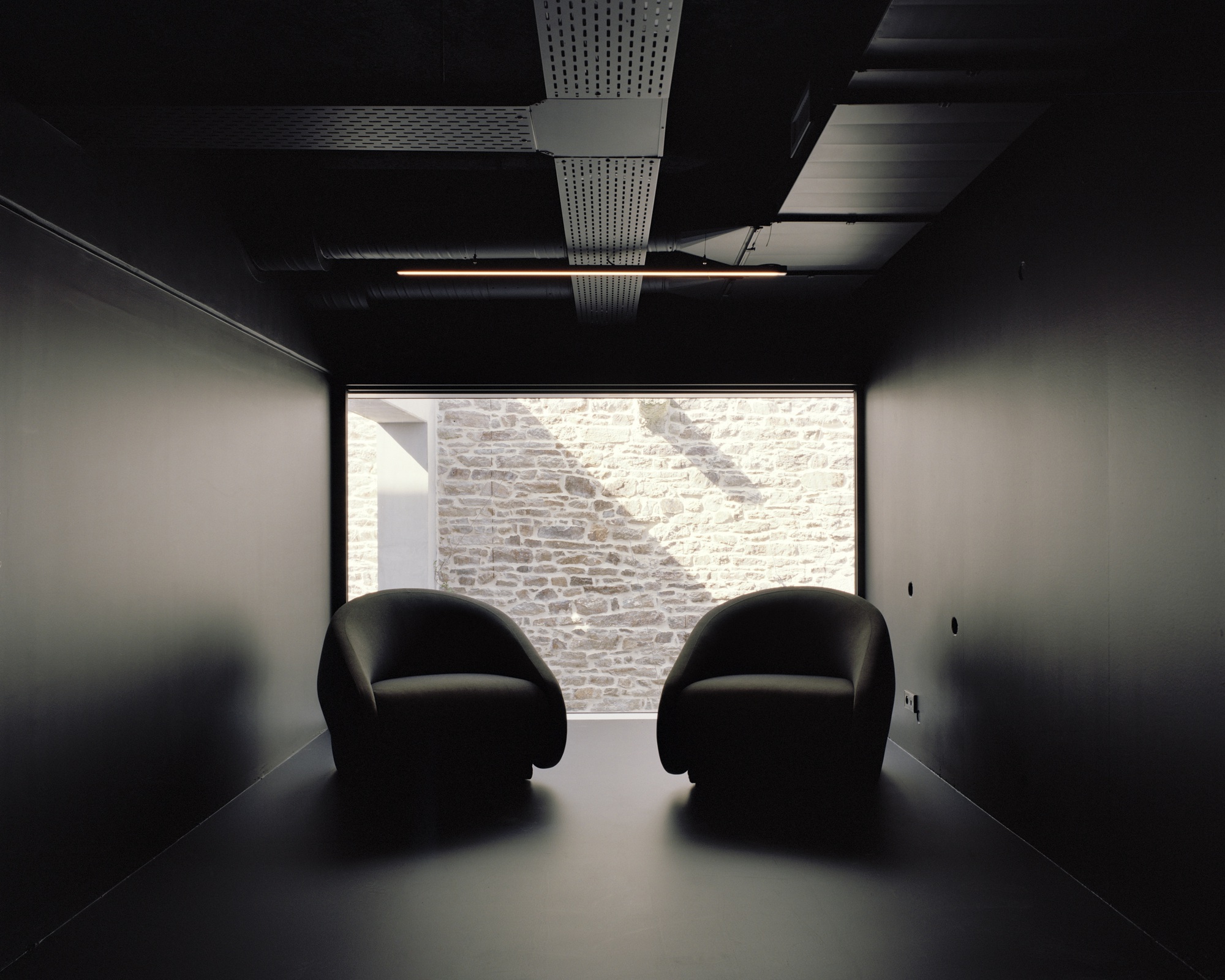 Dark room with two rounded chairs facing a window framing a sunlit stone wall