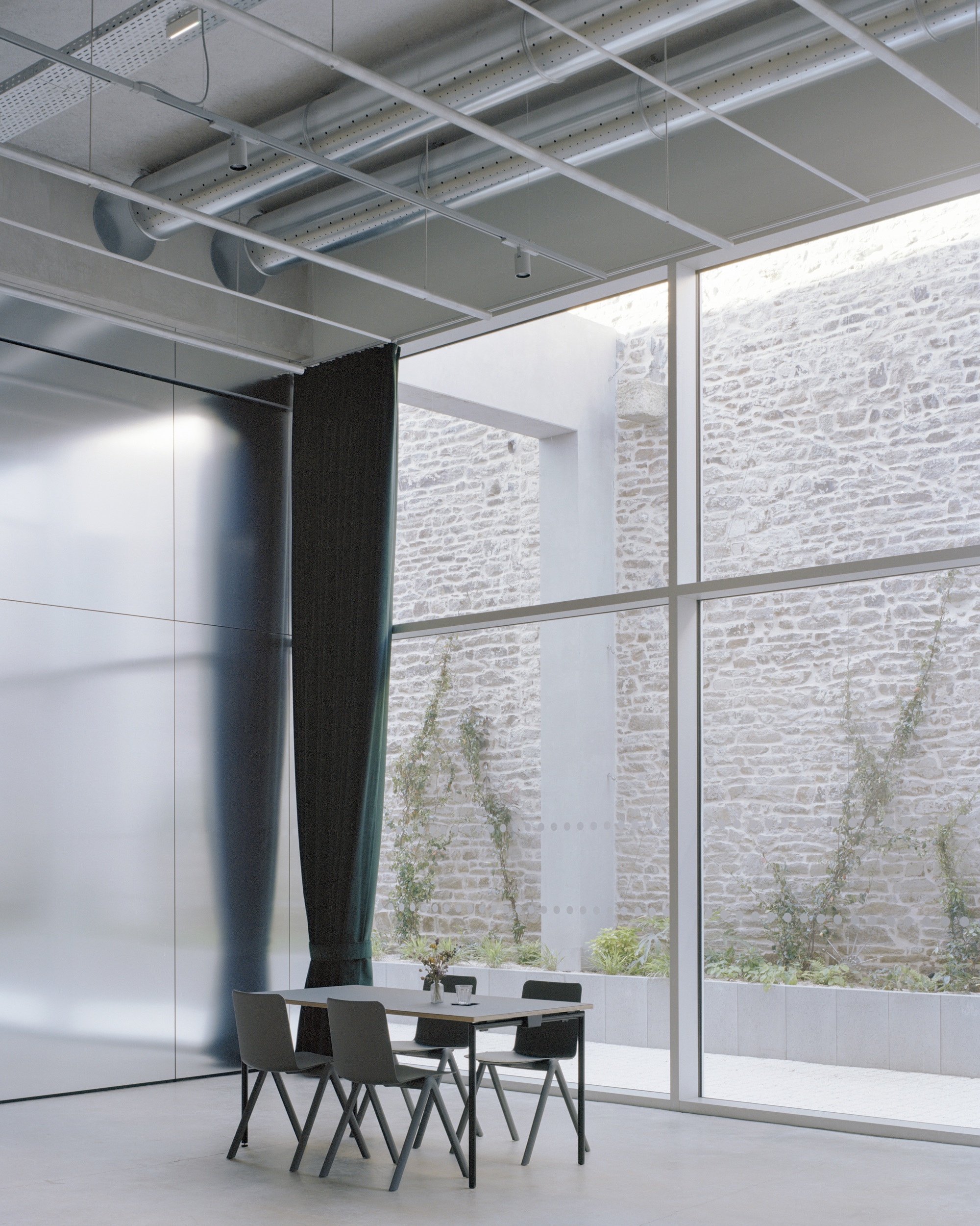 Double-height glazed wall revealing a courtyard with planted saplings against a stone masonry facade