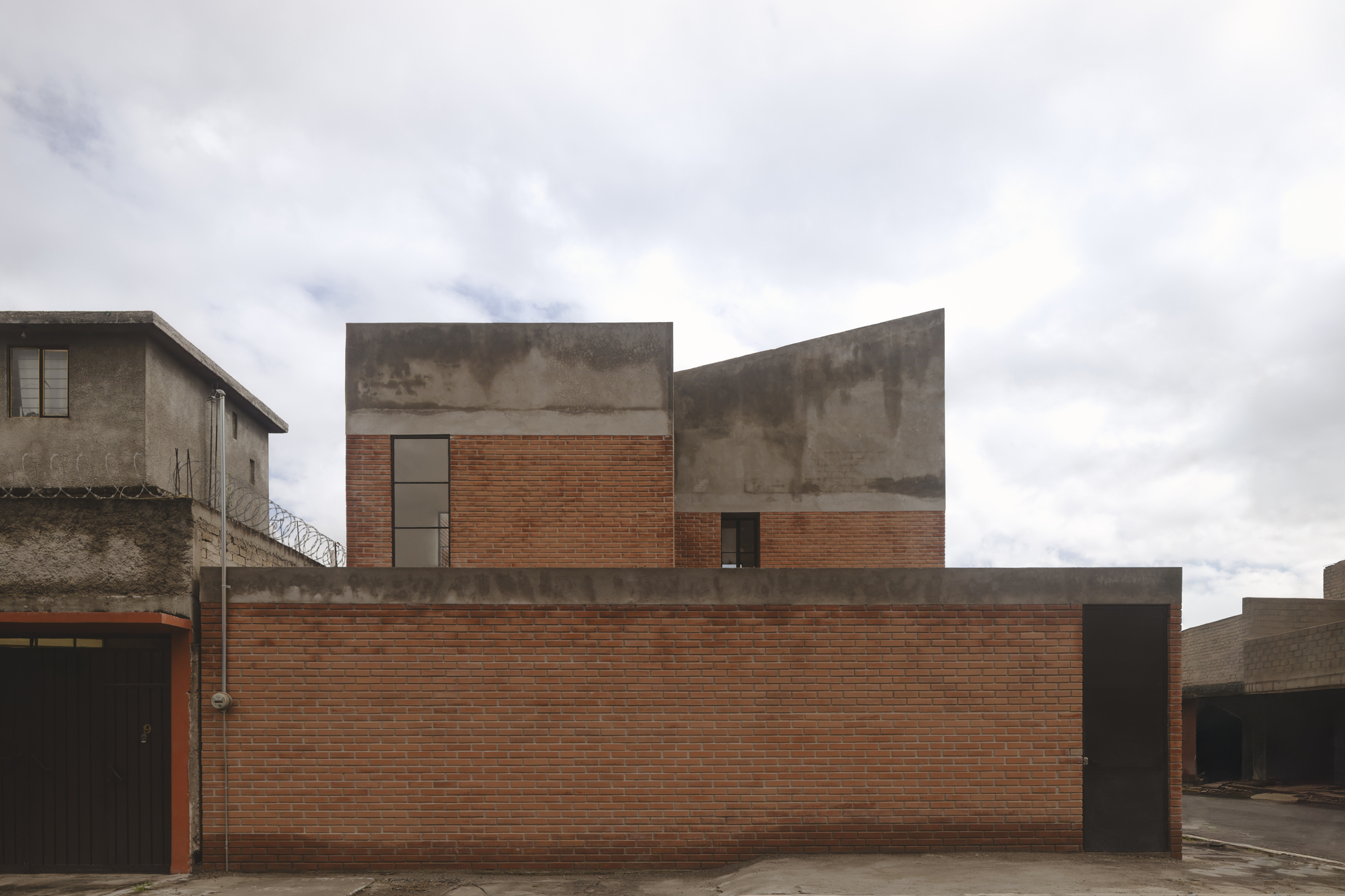 Street facade with stepped concrete volumes rising above a continuous red brick wall under cloudy sky