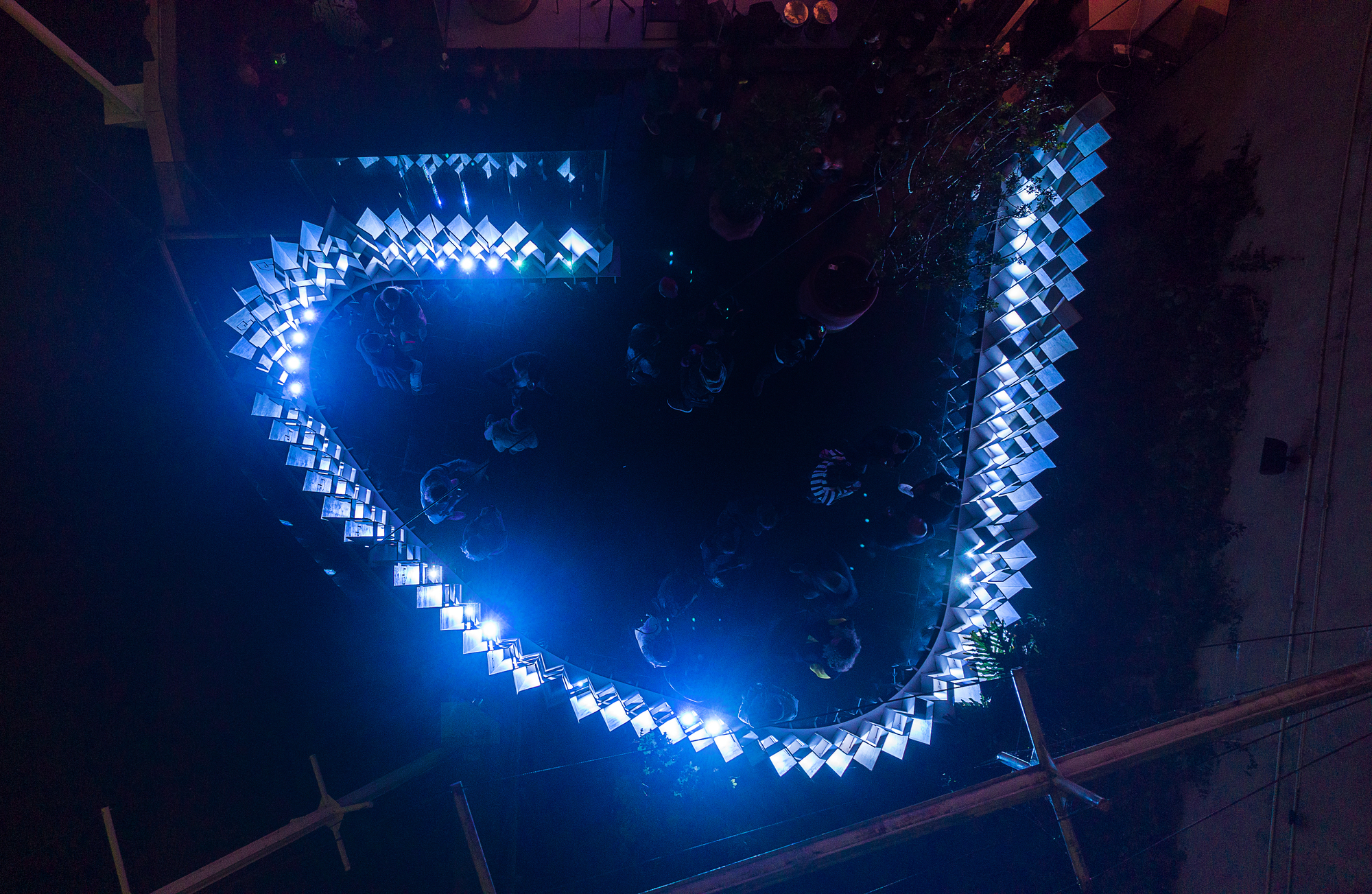 Overhead view of the horseshoe-shaped glowing pavilion with visitors moving through the courtyard
