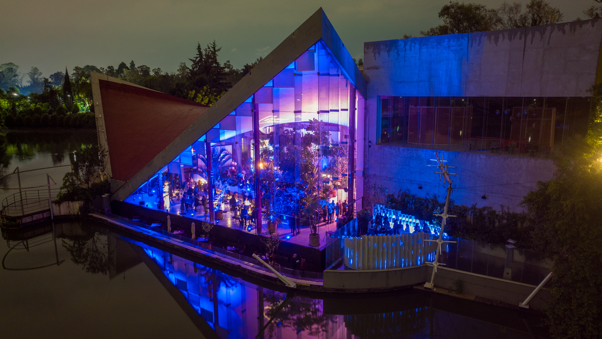 Glass pavilion illuminated in blue and purple light at dusk with reflection in the water