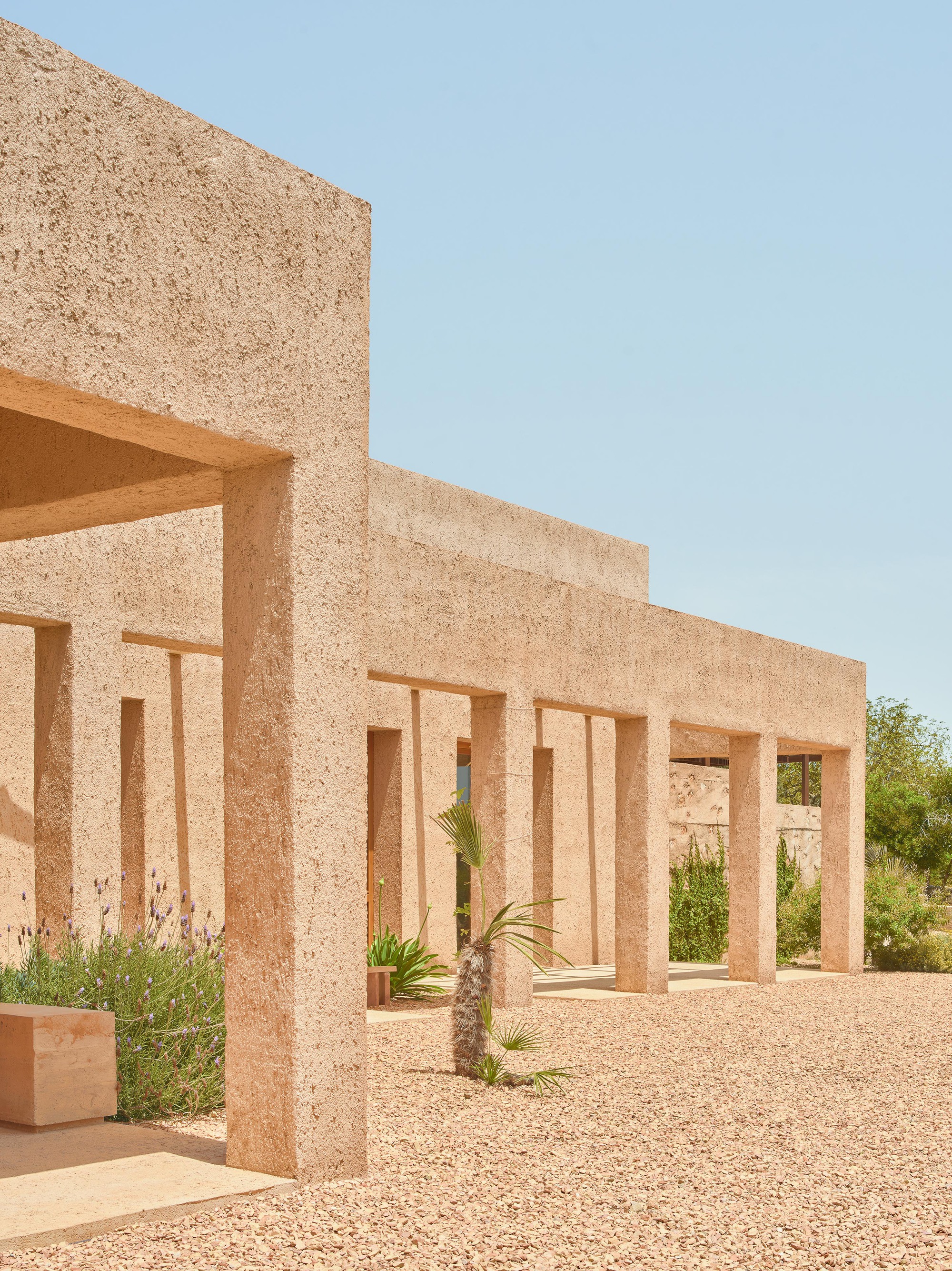 Textured aggregate block colonnade framing a gravel courtyard with planted palms under clear sky