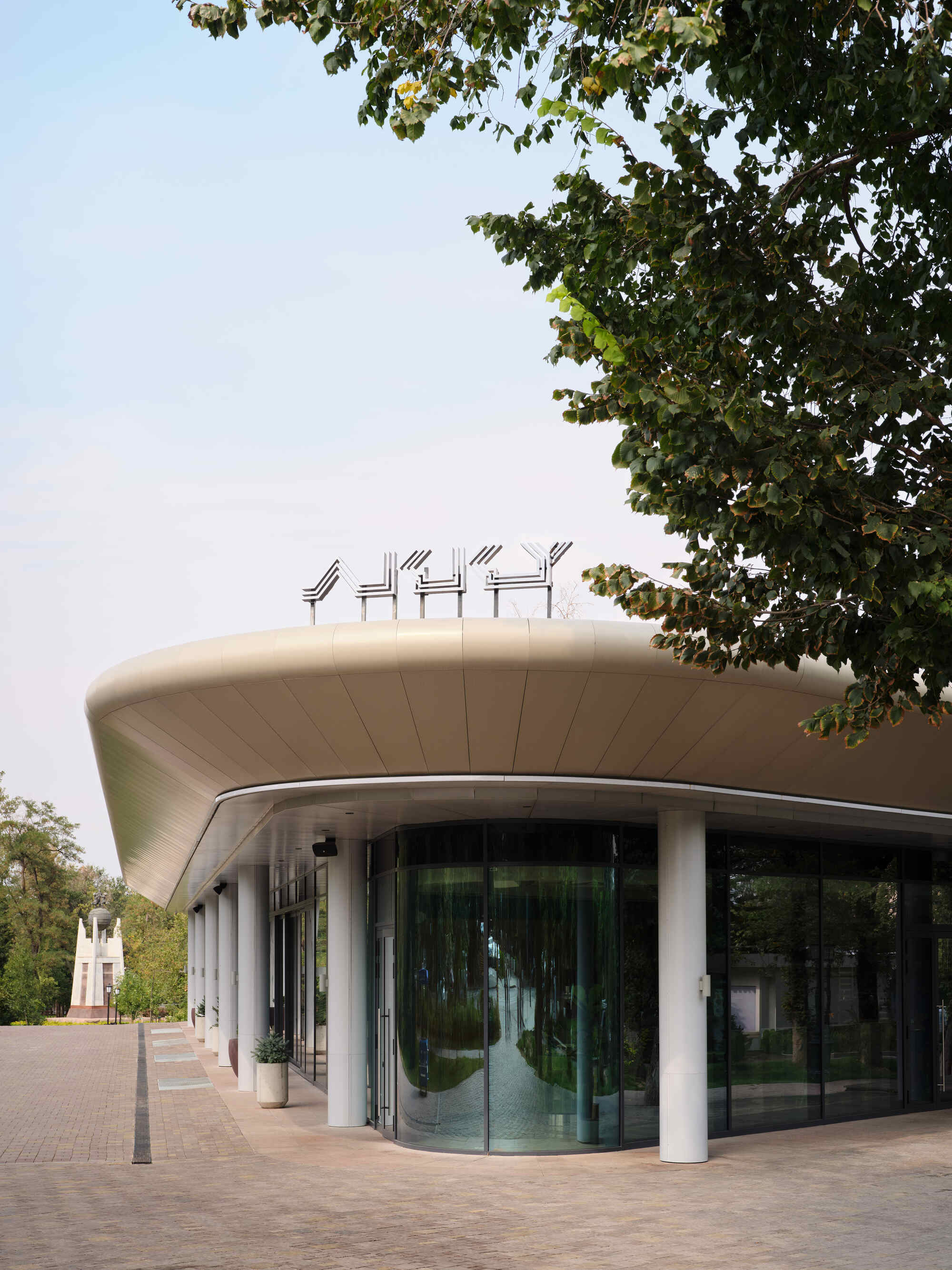 Curved facade with white columns and glazing beneath an overhanging roof framed by tree canopy