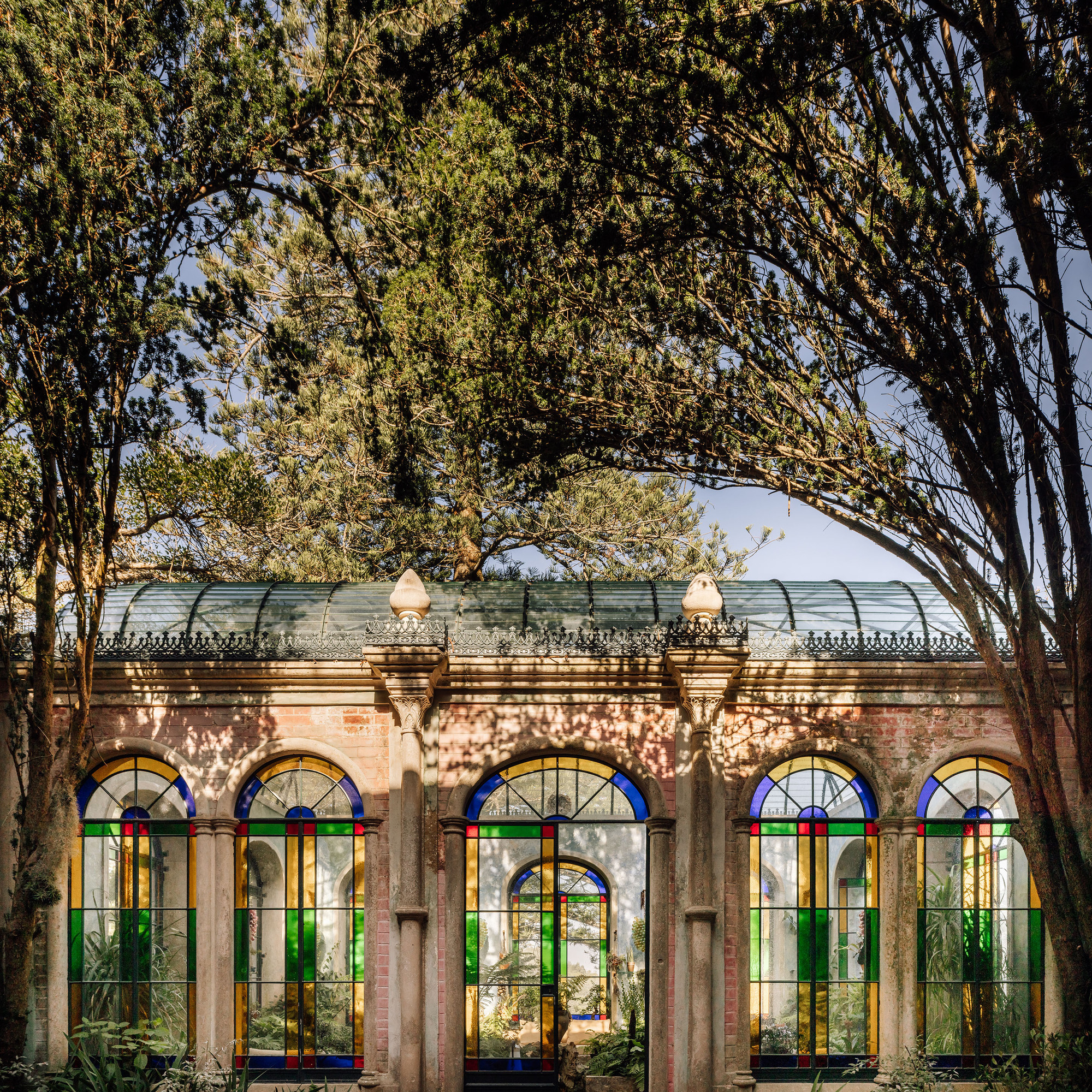 Row of arched windows with colorful stained glass beneath a barrel vault roof under mature tree canopy