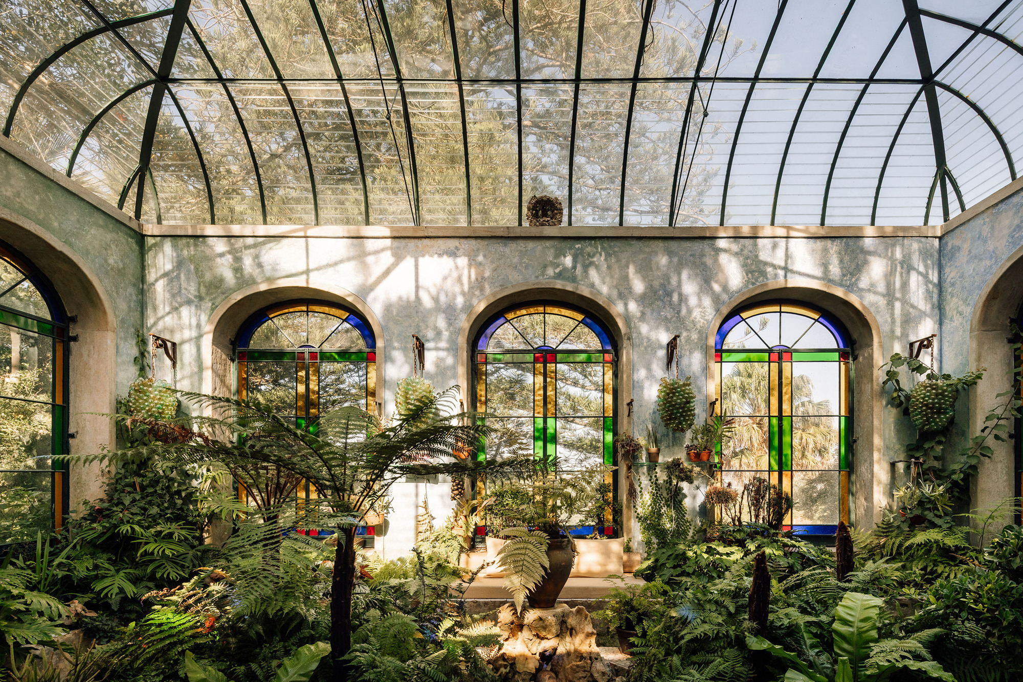 Interior conservatory with arched glass roof and stained glass windows framing potted plants and stone fountain