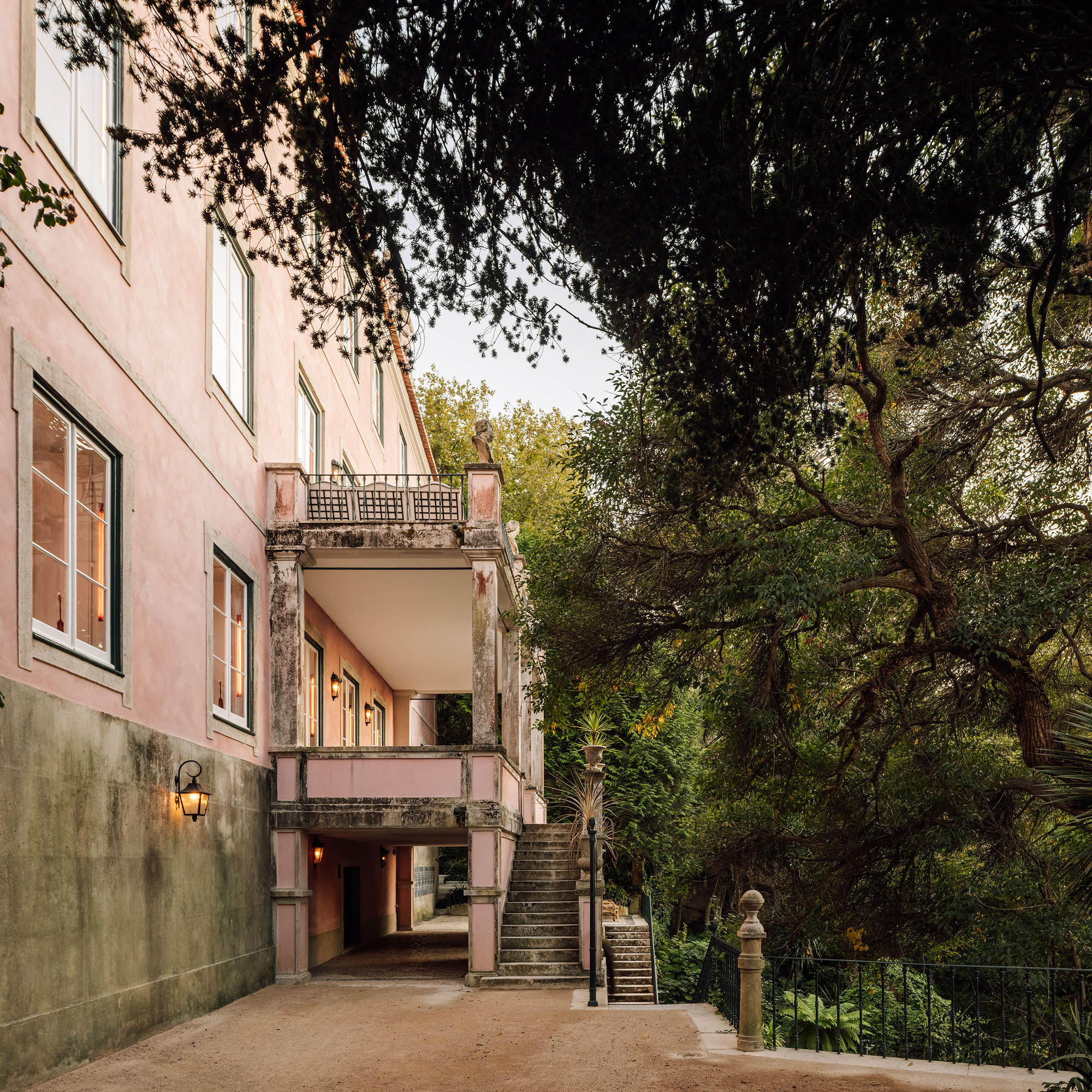 Covered exterior passage with twin staircases and arched openings framed by mature trees at dusk