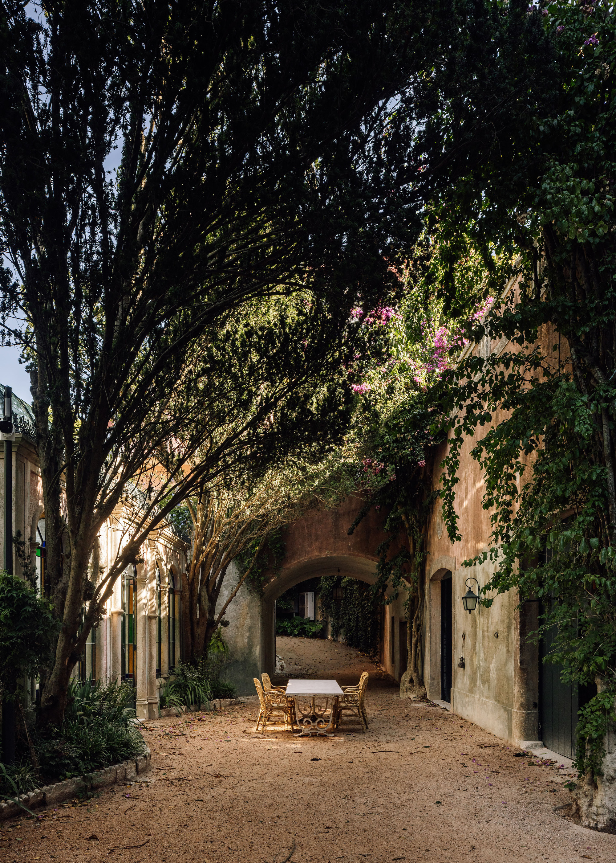 Gravel courtyard with wicker furniture under vine-covered arches and overhanging tree canopy