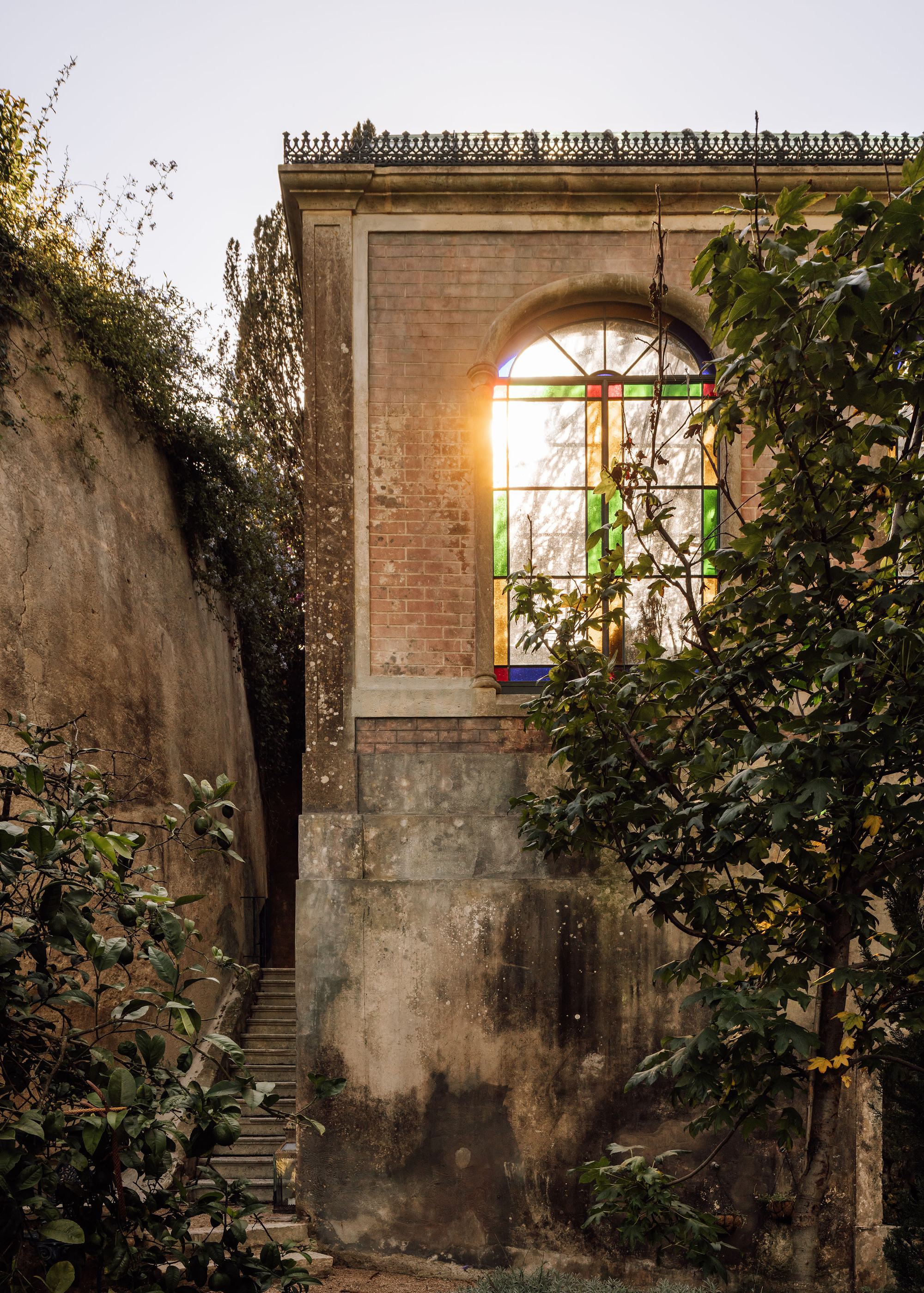 Weathered brick pavilion with arched stained glass window illuminated at dusk amid climbing vegetation