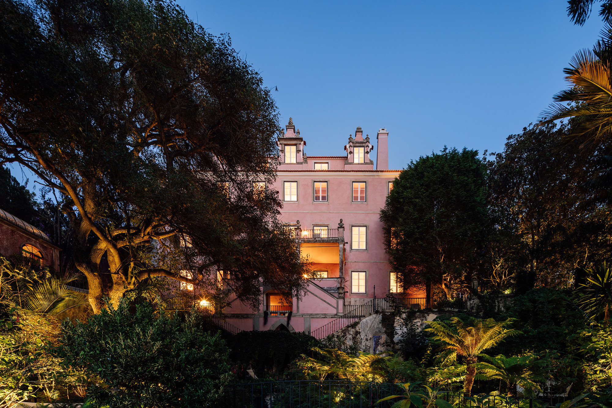 Pink rendered facade with symmetrical chimneys framed by palms and trees at dusk