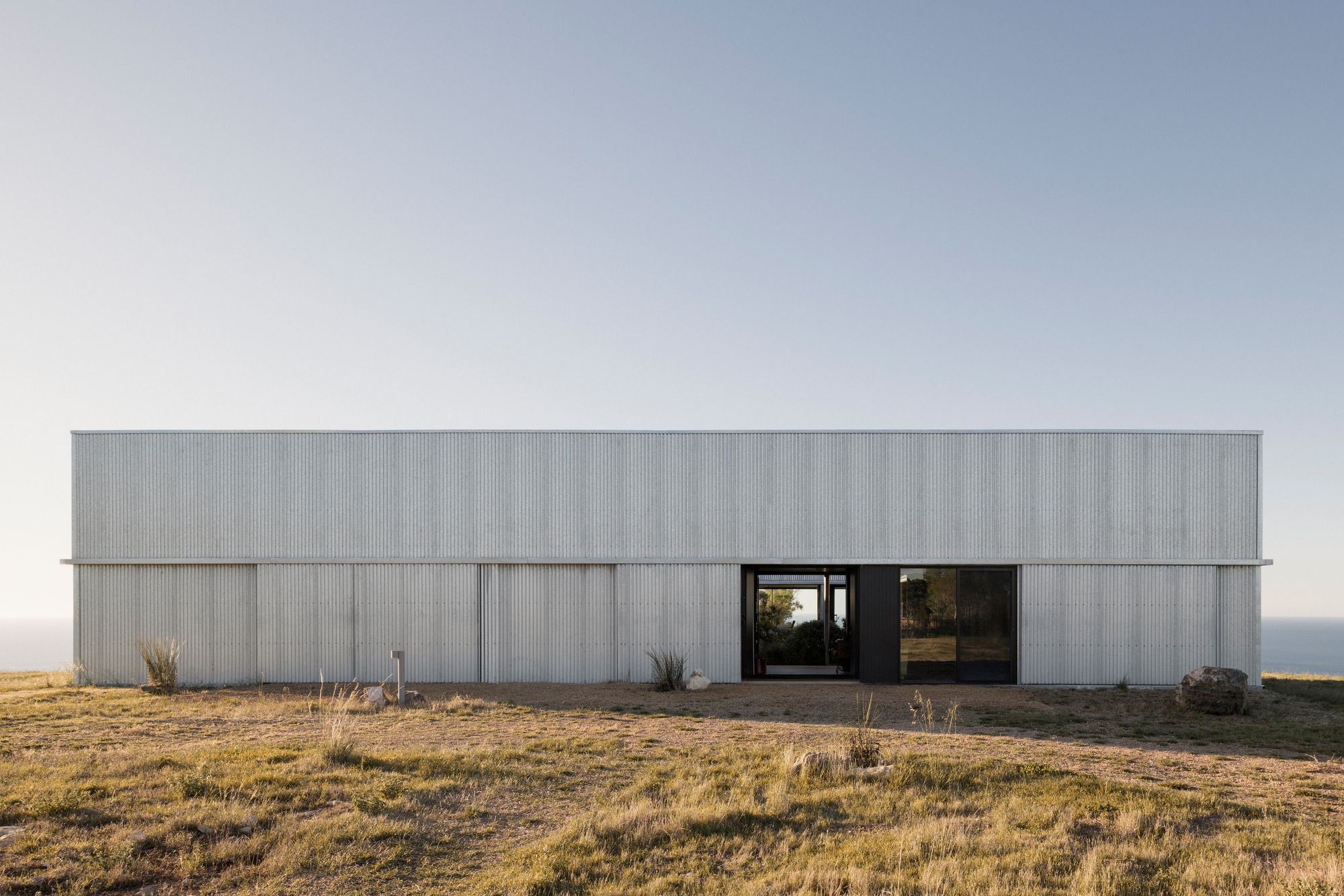 Long horizontal volume clad in vertical corrugated metal with central recessed entry at dusk