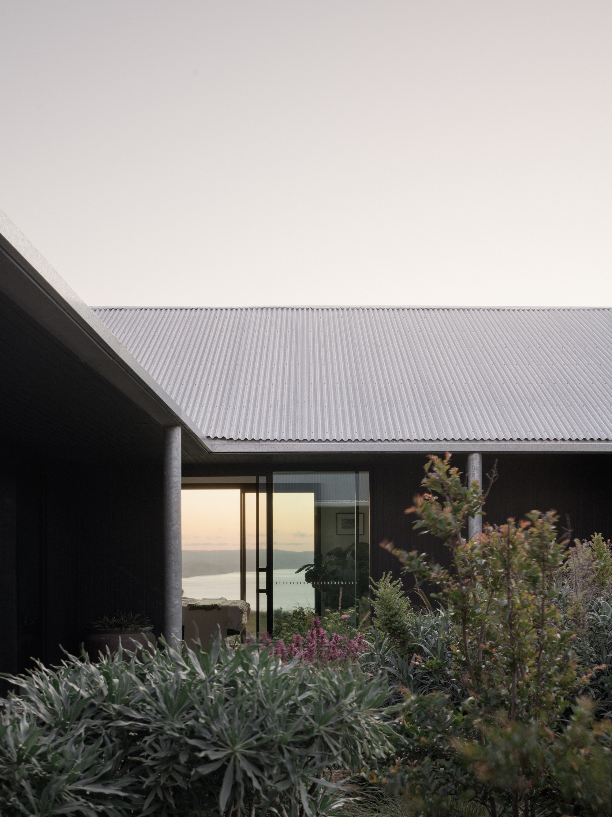 Corner of the corrugated metal roof overhang above glazed walls and planted beds at sunset