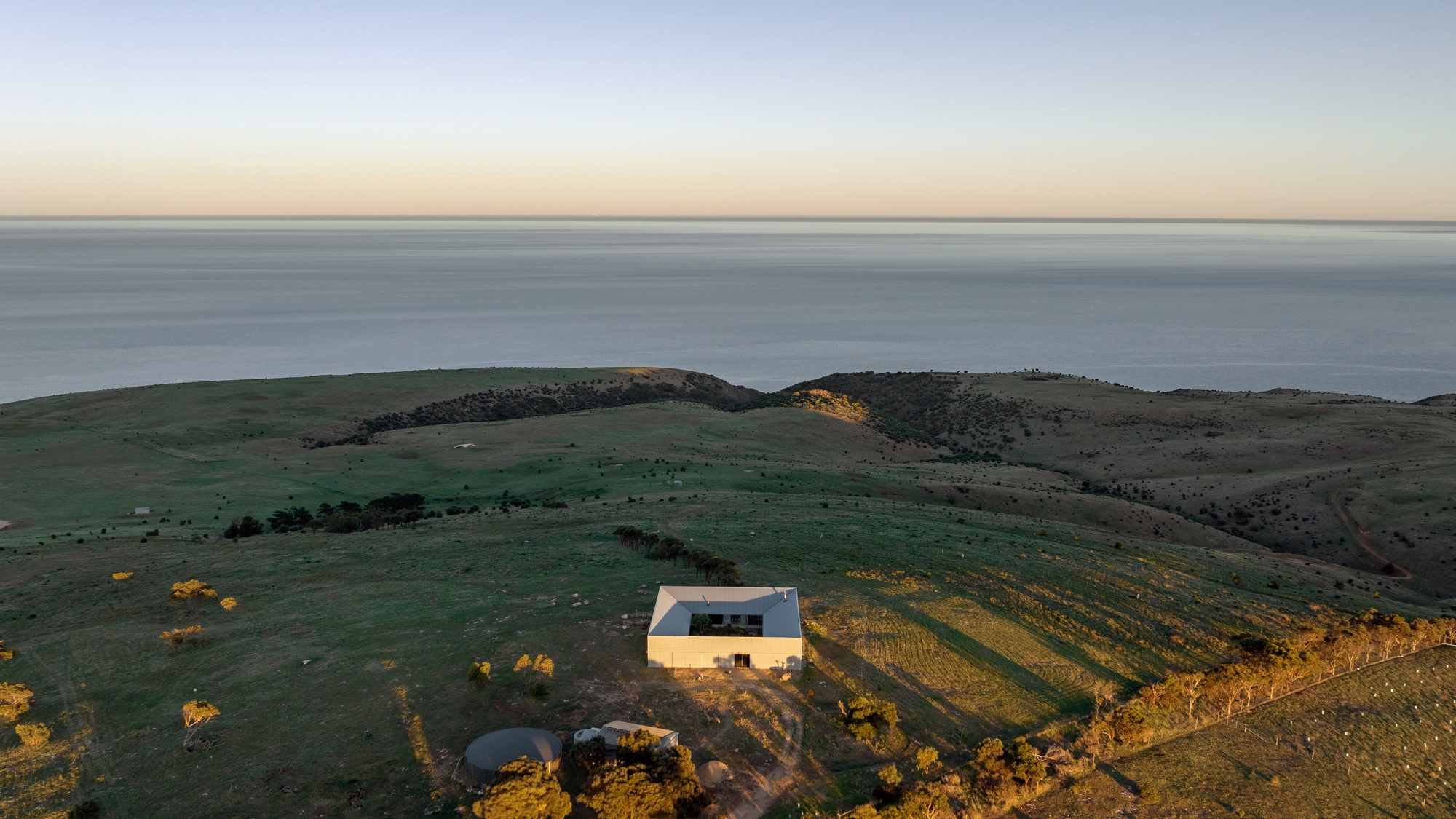 Aerial view of a white rectangular building on sloped farmland overlooking the ocean at dusk