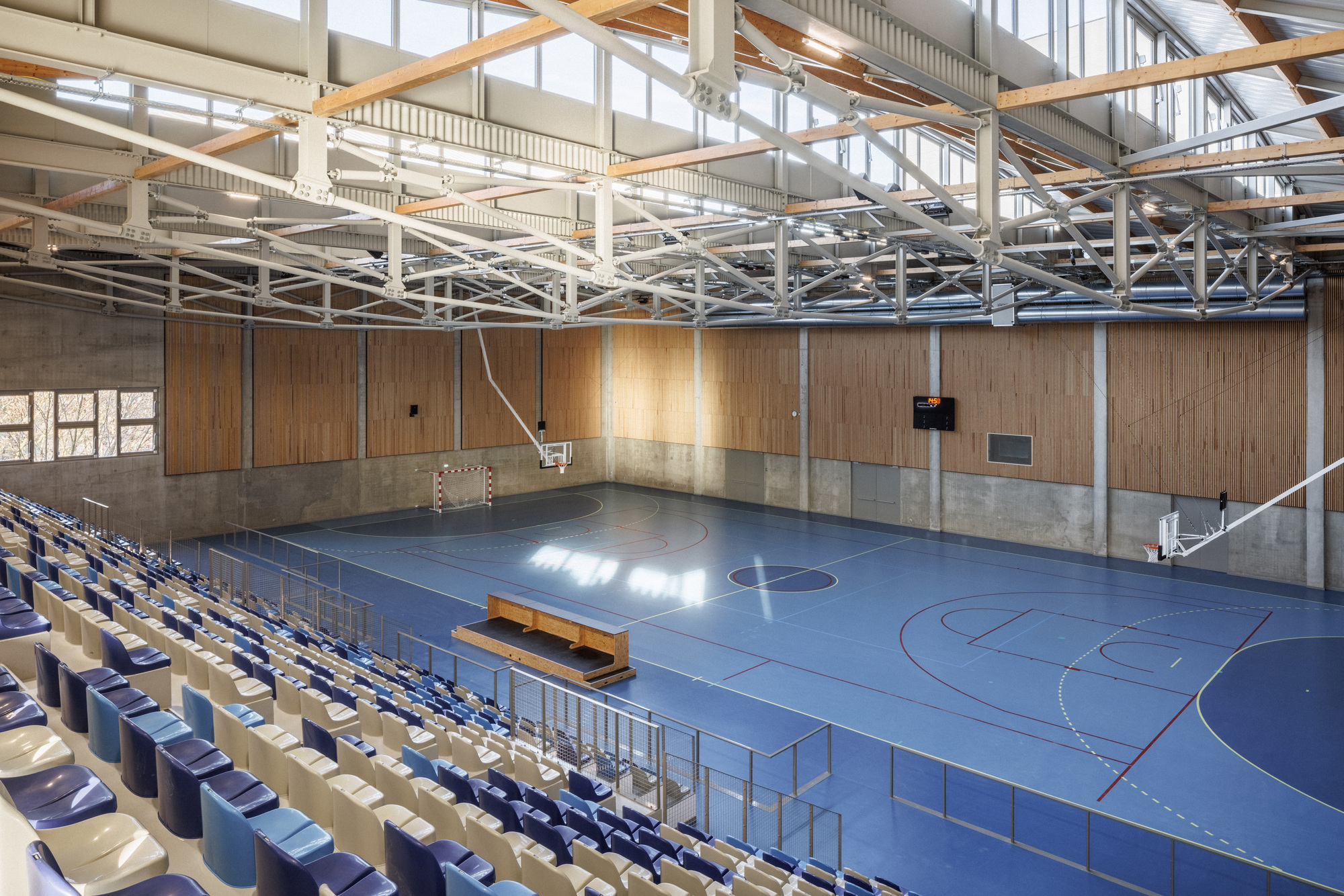 Interior view of the sports hall showing exposed timber and steel trusses above blue flooring and bleachers