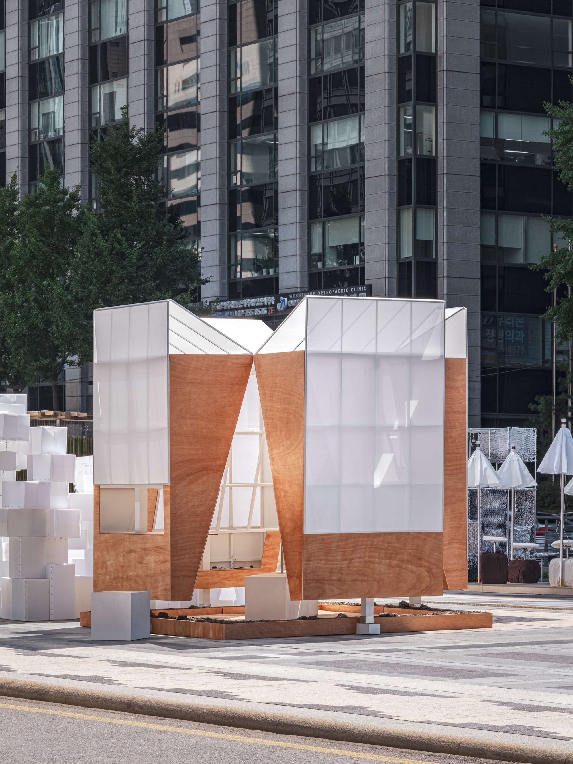 Timber and translucent panel pavilion with triangular openings framing views of residential towers behind