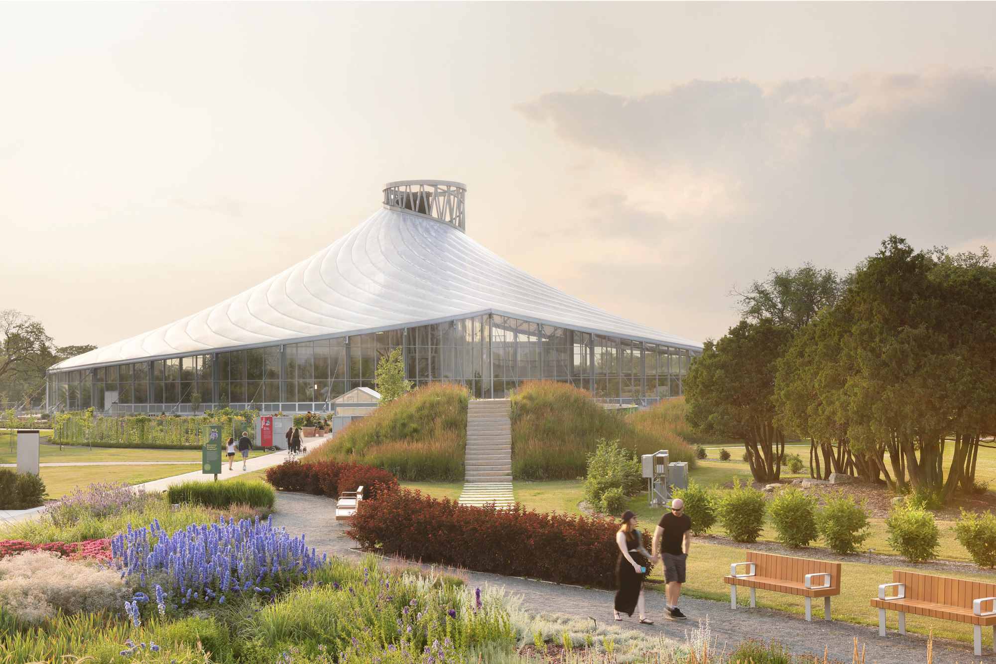 White tensile roof rising to a central lantern above a glass pavilion surrounded by planted beds with visitors