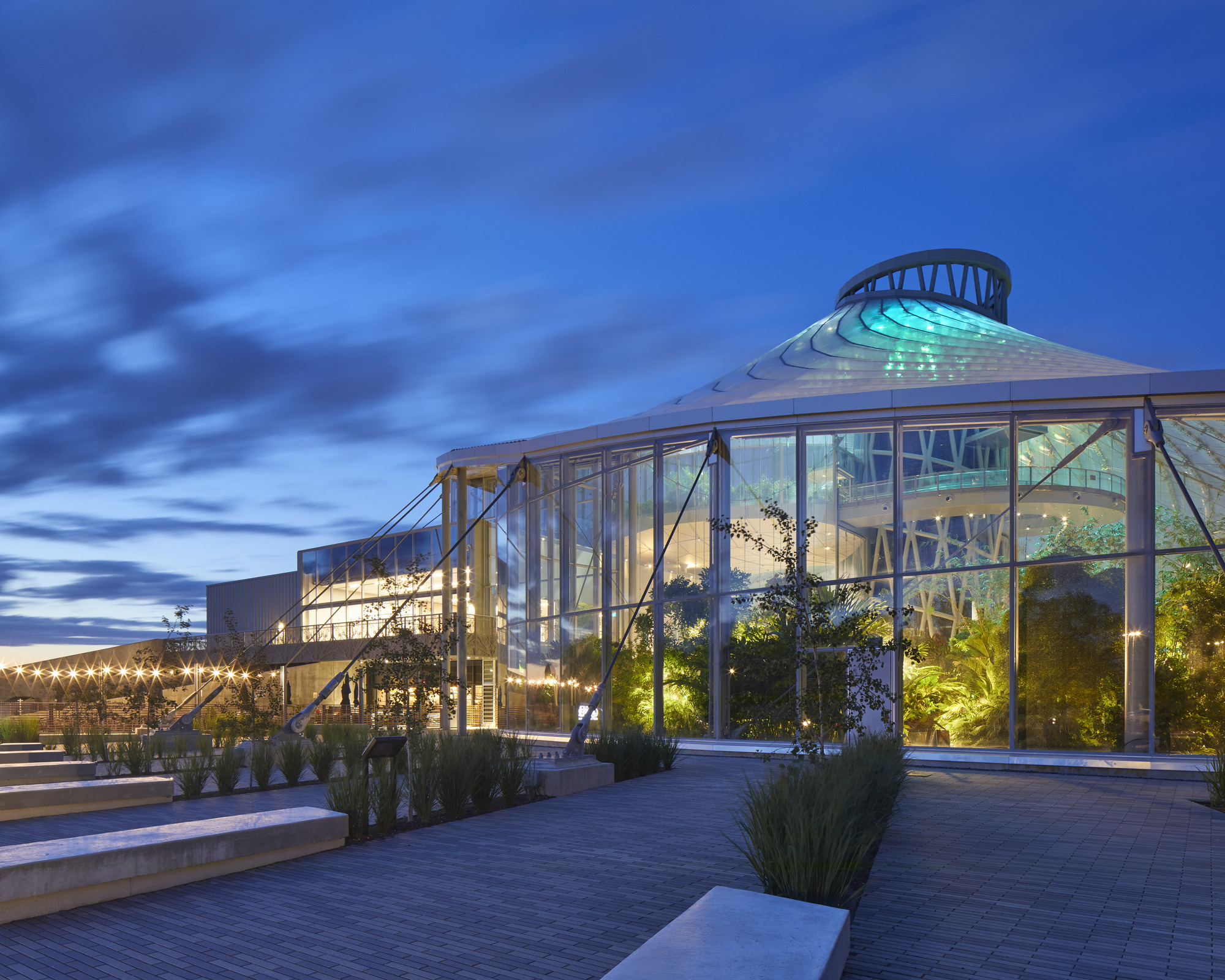 Glass conservatory volume with exposed steel structure and central dome illuminated against a blue evening sky