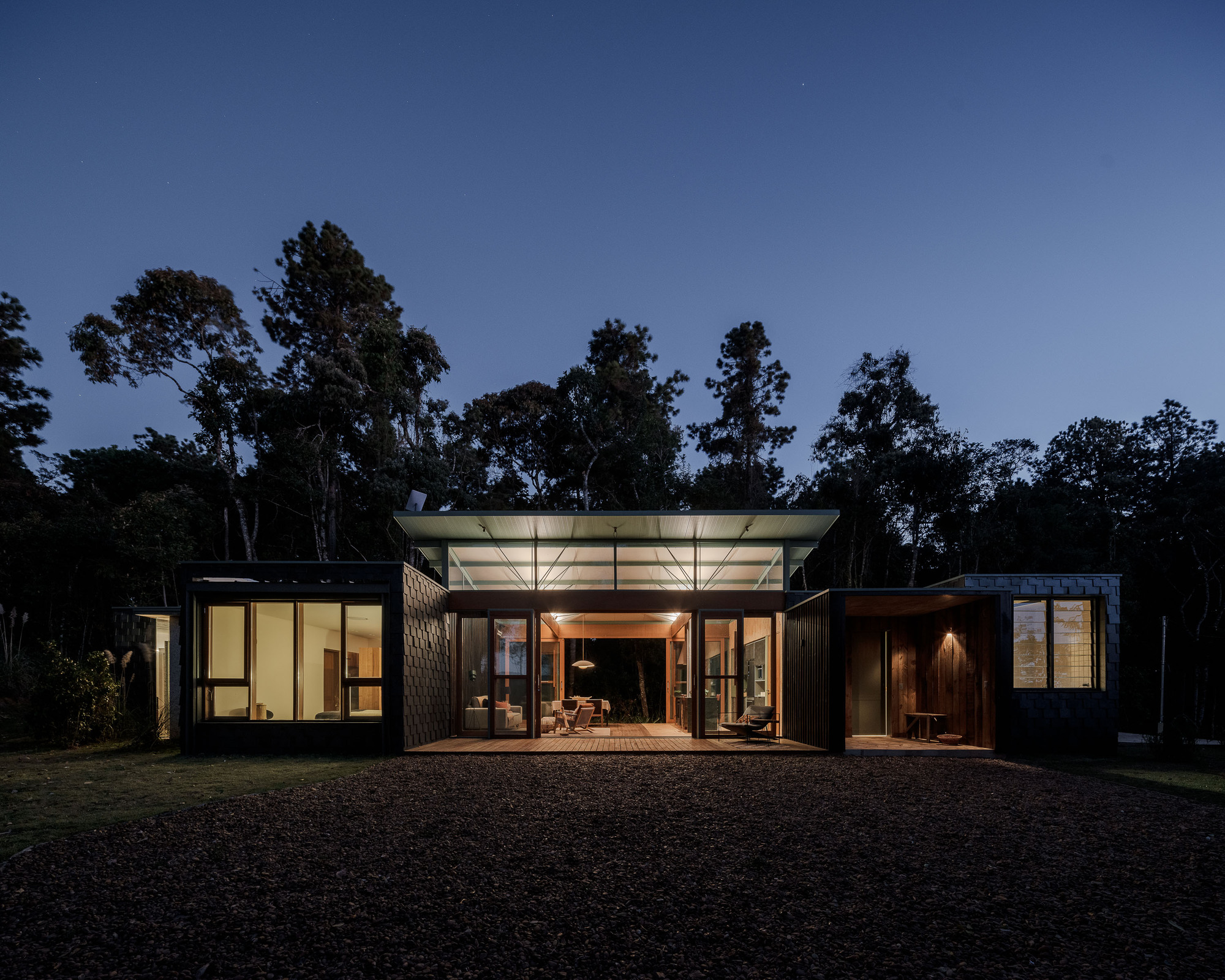 Evening view of the flat-roofed residence with illuminated interiors framed by glass walls and dark cladding panels