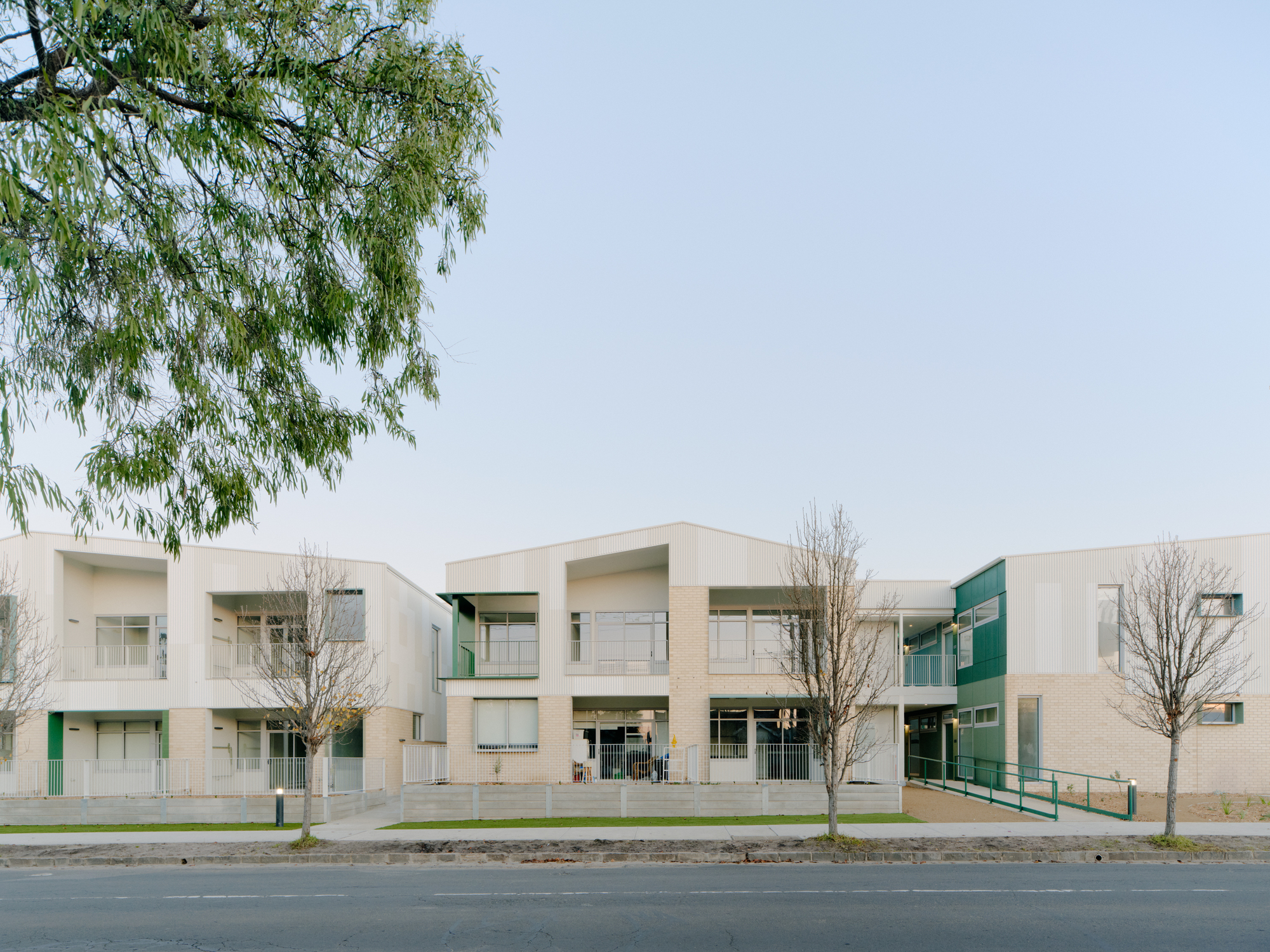 Street view of the two-story facade with white corrugated cladding, recessed balconies and bare winter trees