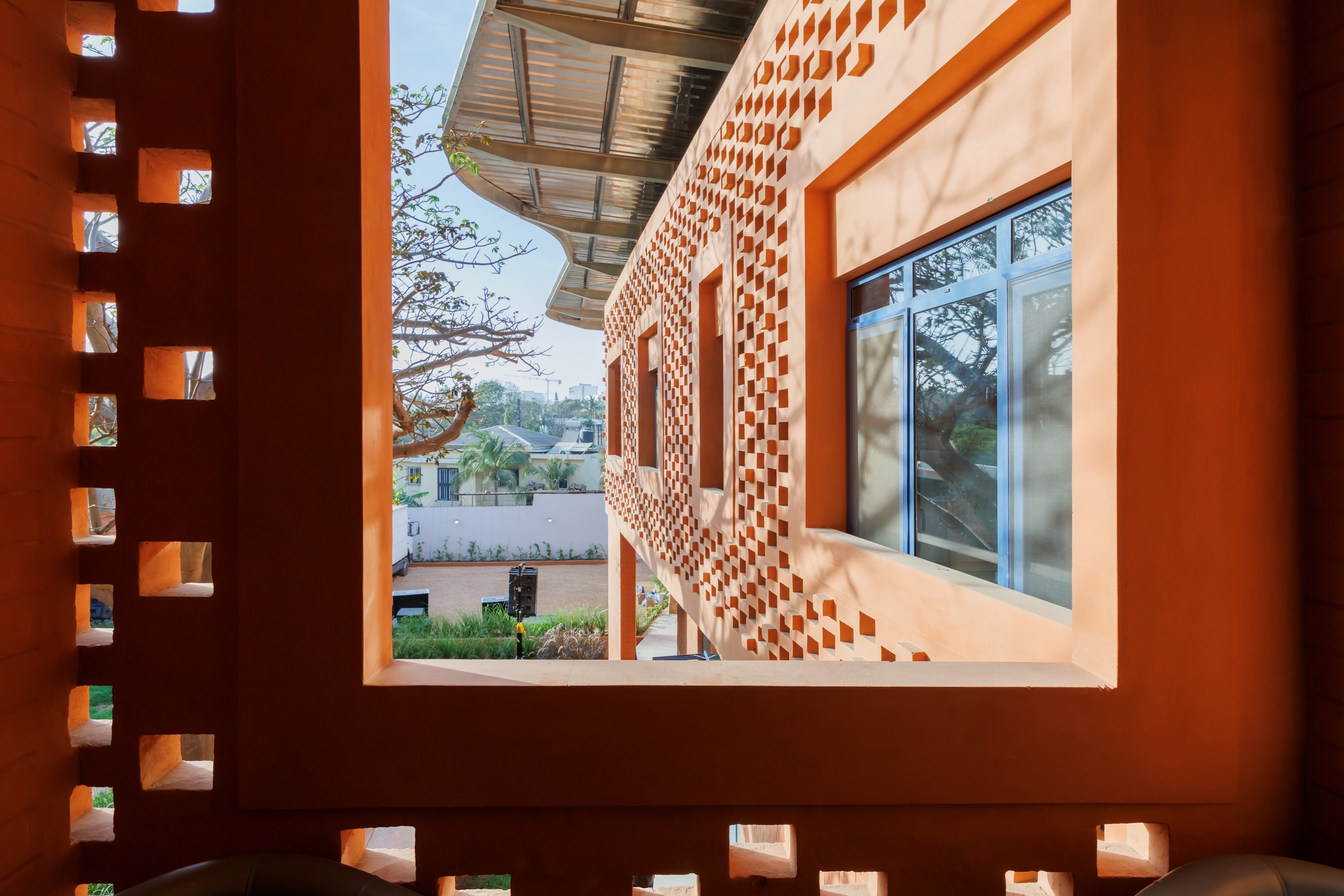 View through perforated brick wall opening to courtyard showing curved facade and metal canopy beyond