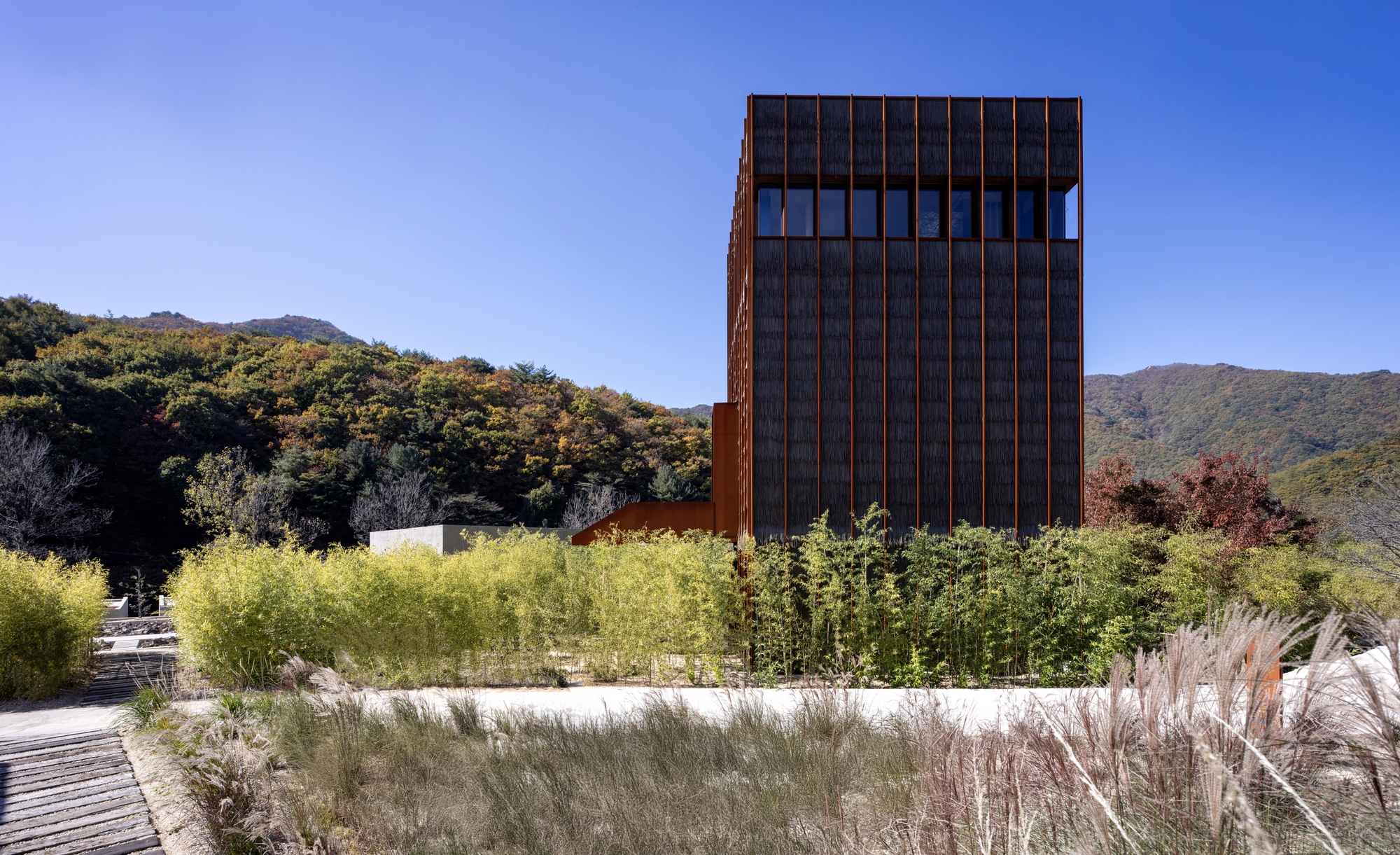 Corten steel tower rising above native grasses and planted beds with forested hills in the distance