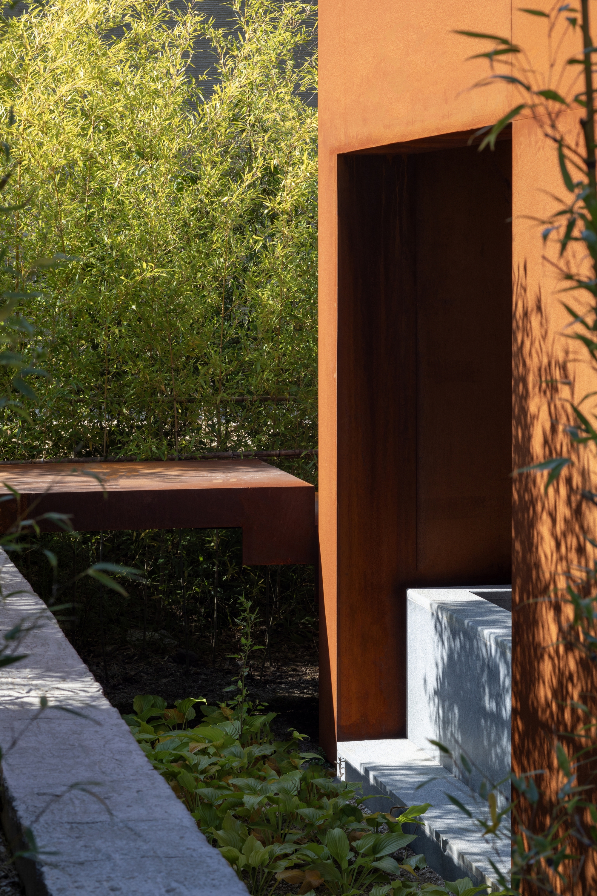 Rusted steel doorway framing bamboo and planted beds with afternoon shadows on the stucco wall