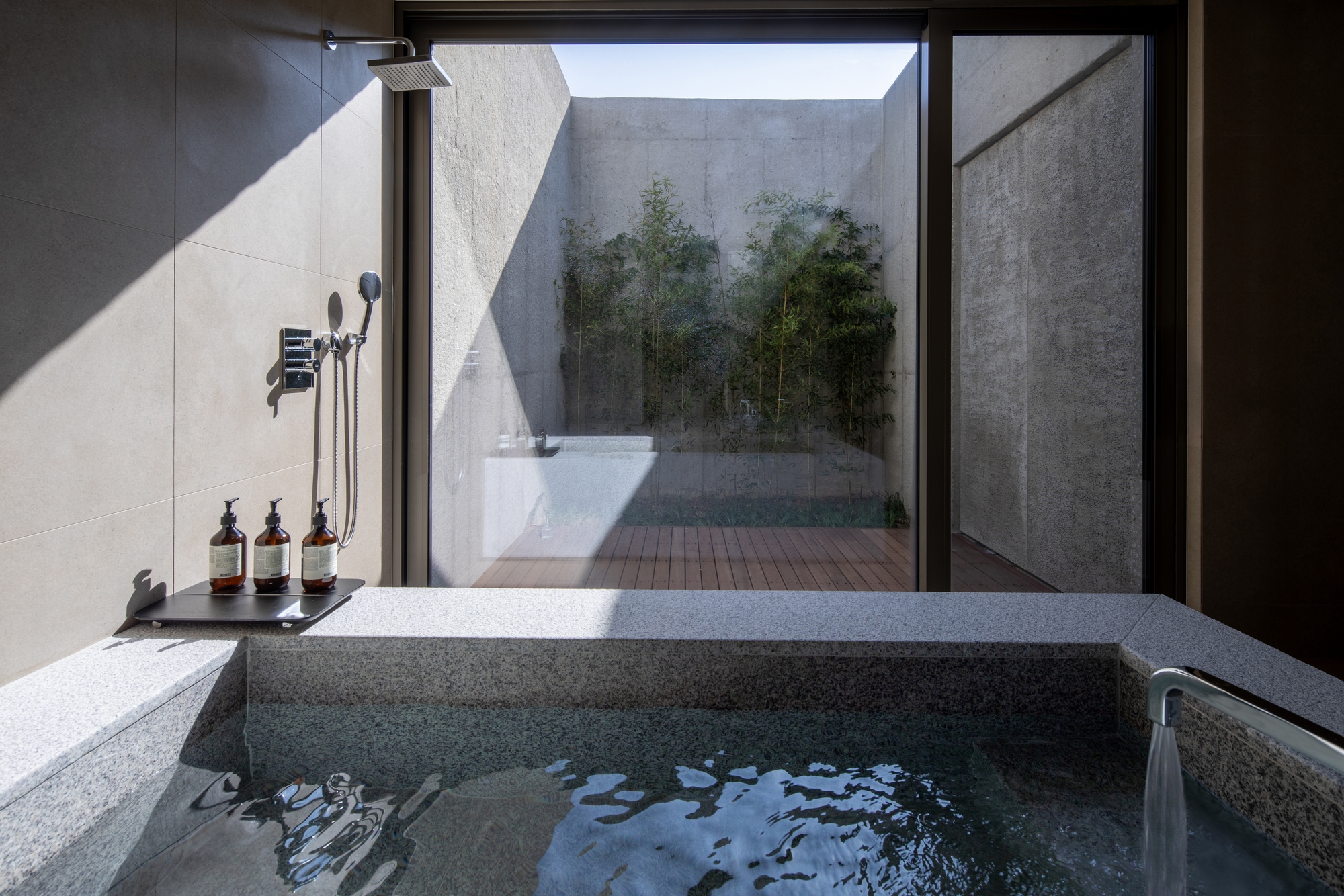 Sunken stone bathtub with rain shower facing a private courtyard with bamboo through floor-to-ceiling glass
