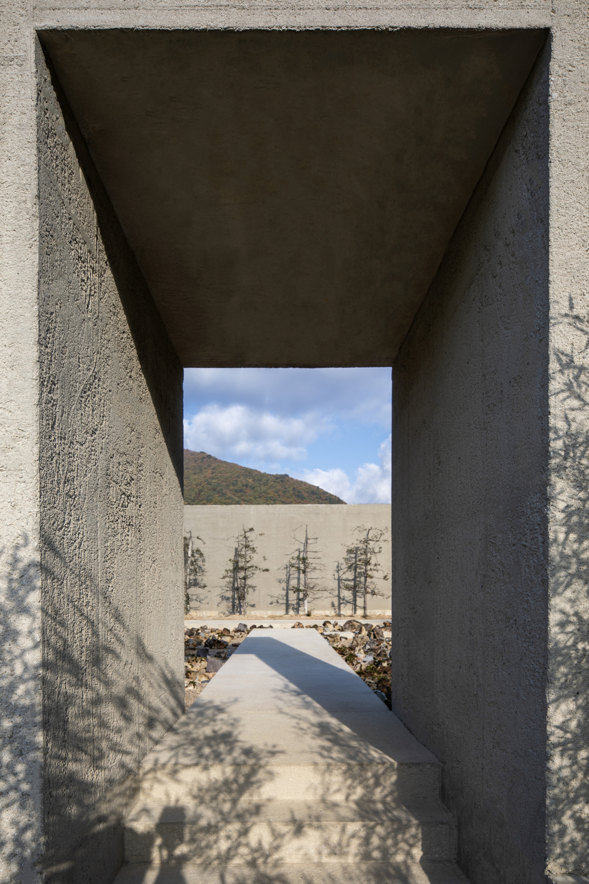 Tapered concrete portal framing a pathway toward young trees and distant hills under a cloudy sky