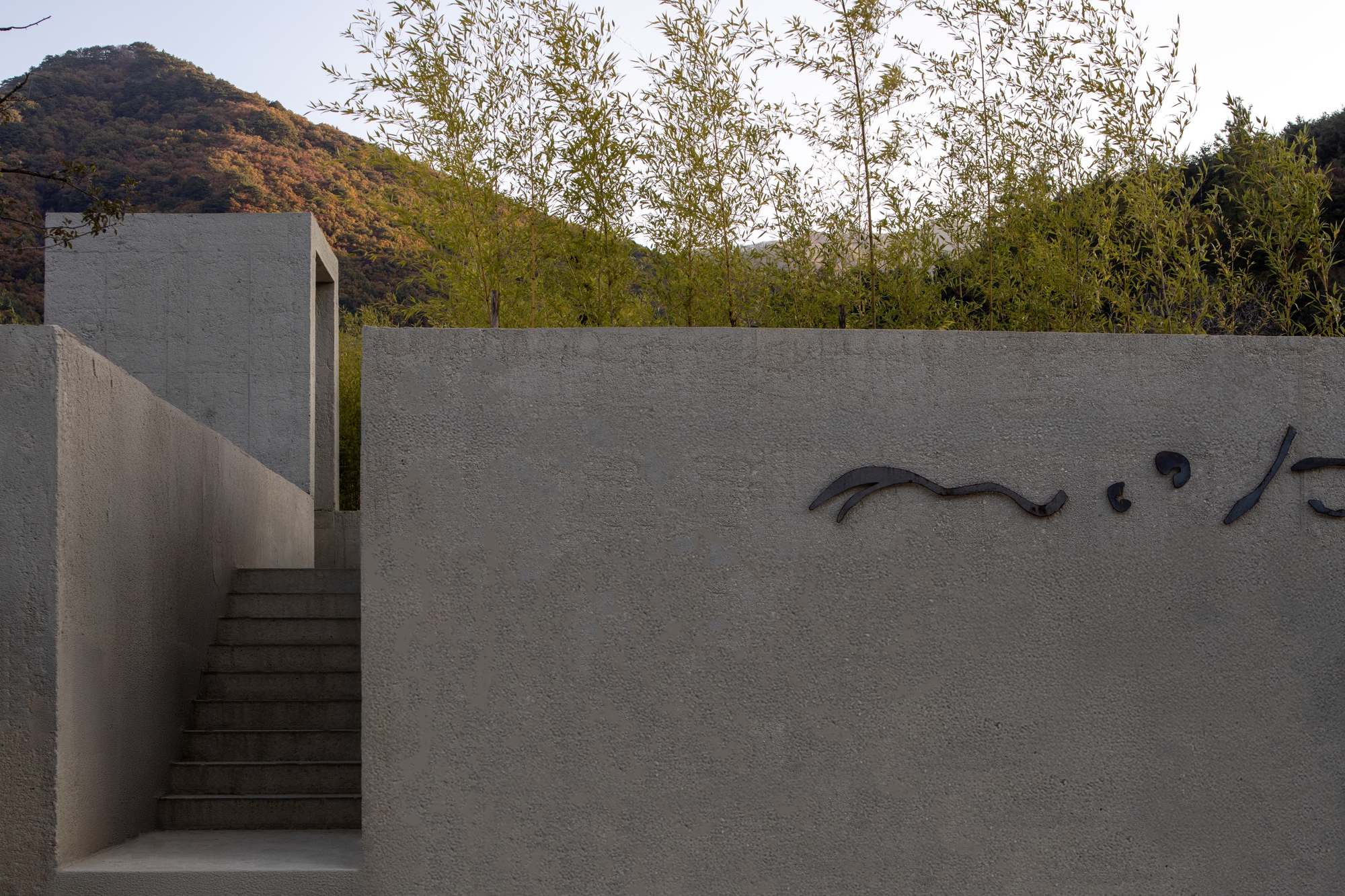 Textured concrete wall with calligraphic metal signage beside an ascending stair at dusk with trees beyond