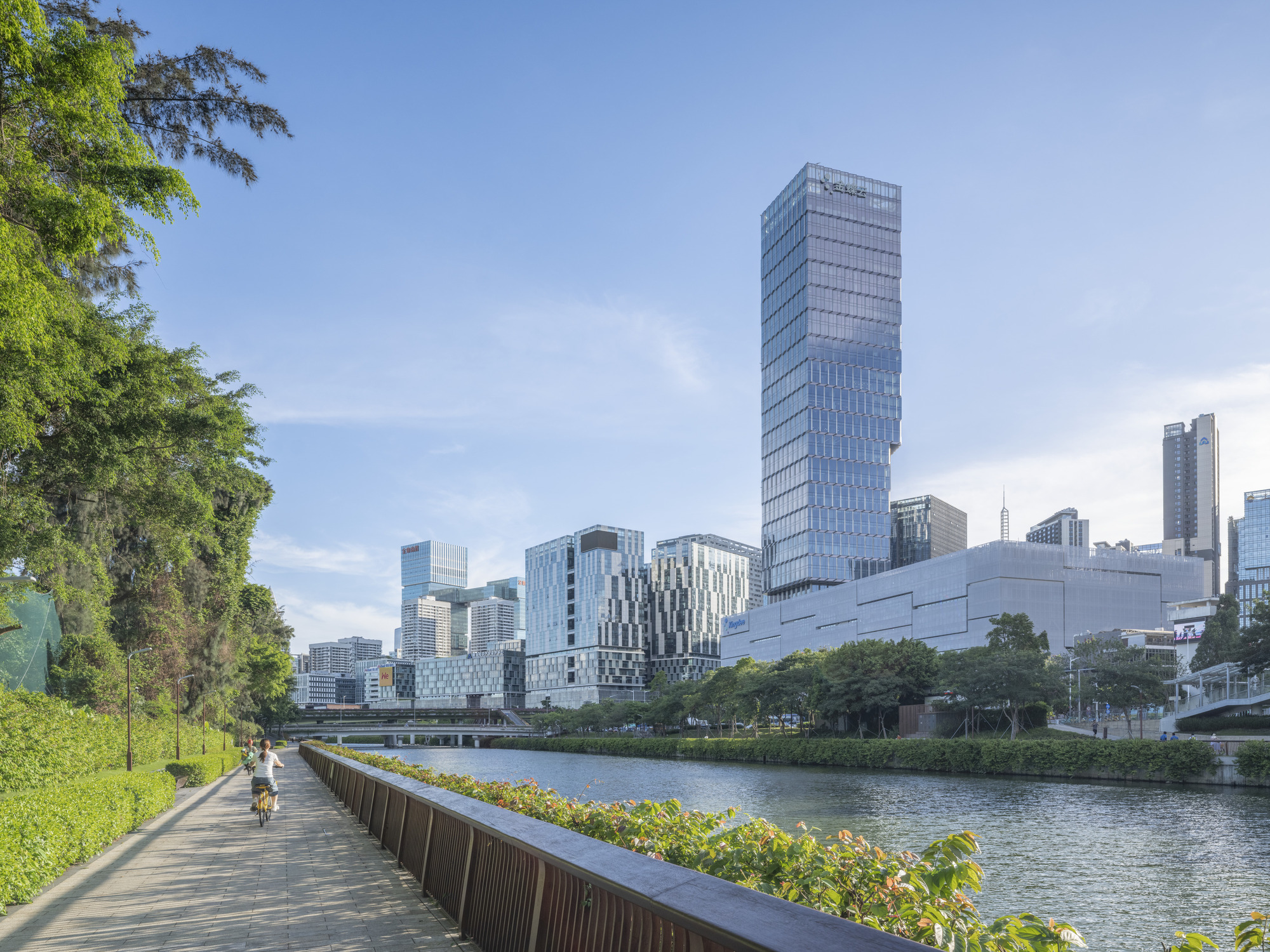 Waterfront pathway with cyclist beneath trees looking toward glazed tower and skyline beyond