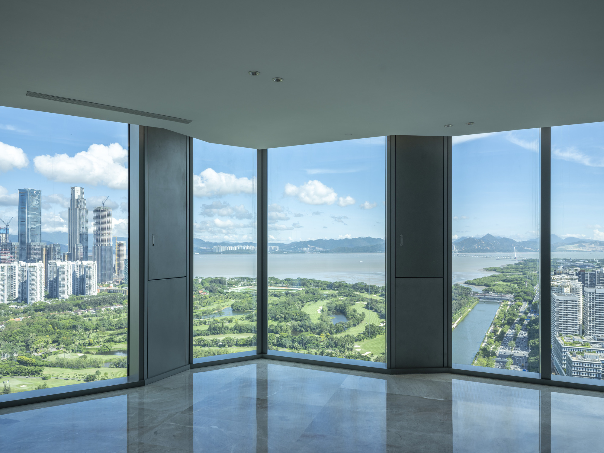 Corner window interior with floor-to-ceiling glass overlooking a golf course and waterfront on a cloudy day