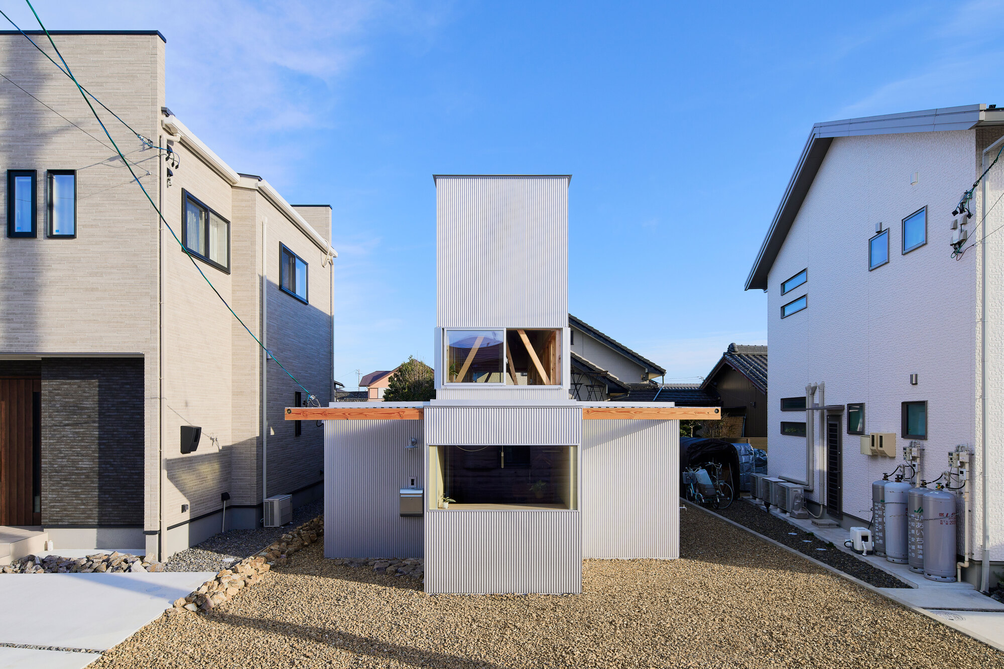 White corrugated metal house with cantilevered upper volume and gravel yard between neighboring buildings