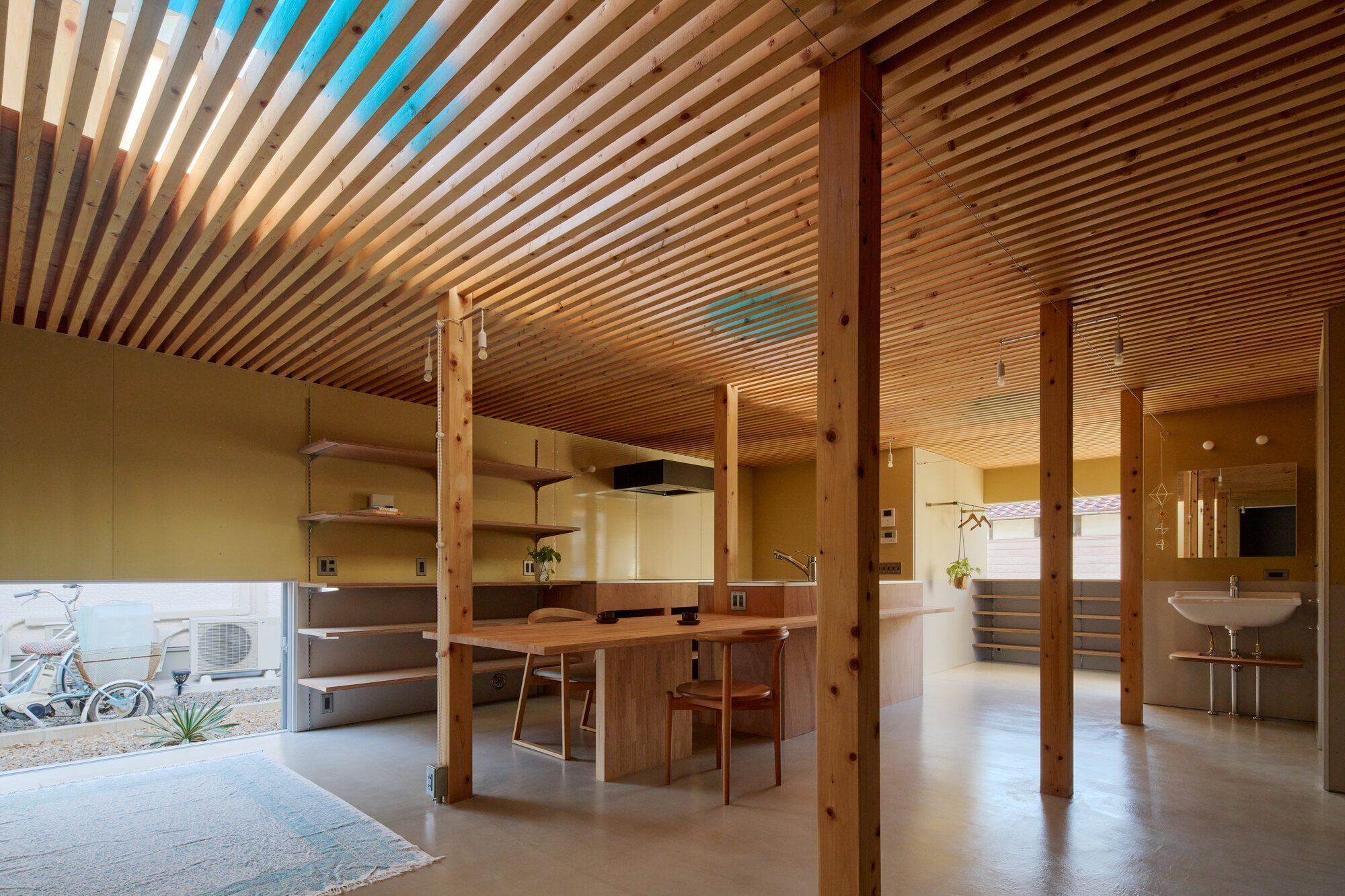 Living and dining area with timber post-and-beam structure, built-in shelving, and slatted ceiling illuminated by pendant lights