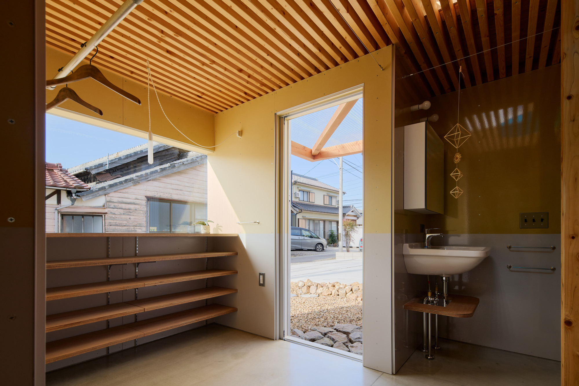Entry corner with timber shelving and wall-mounted sink opening to a gravel courtyard and neighboring houses