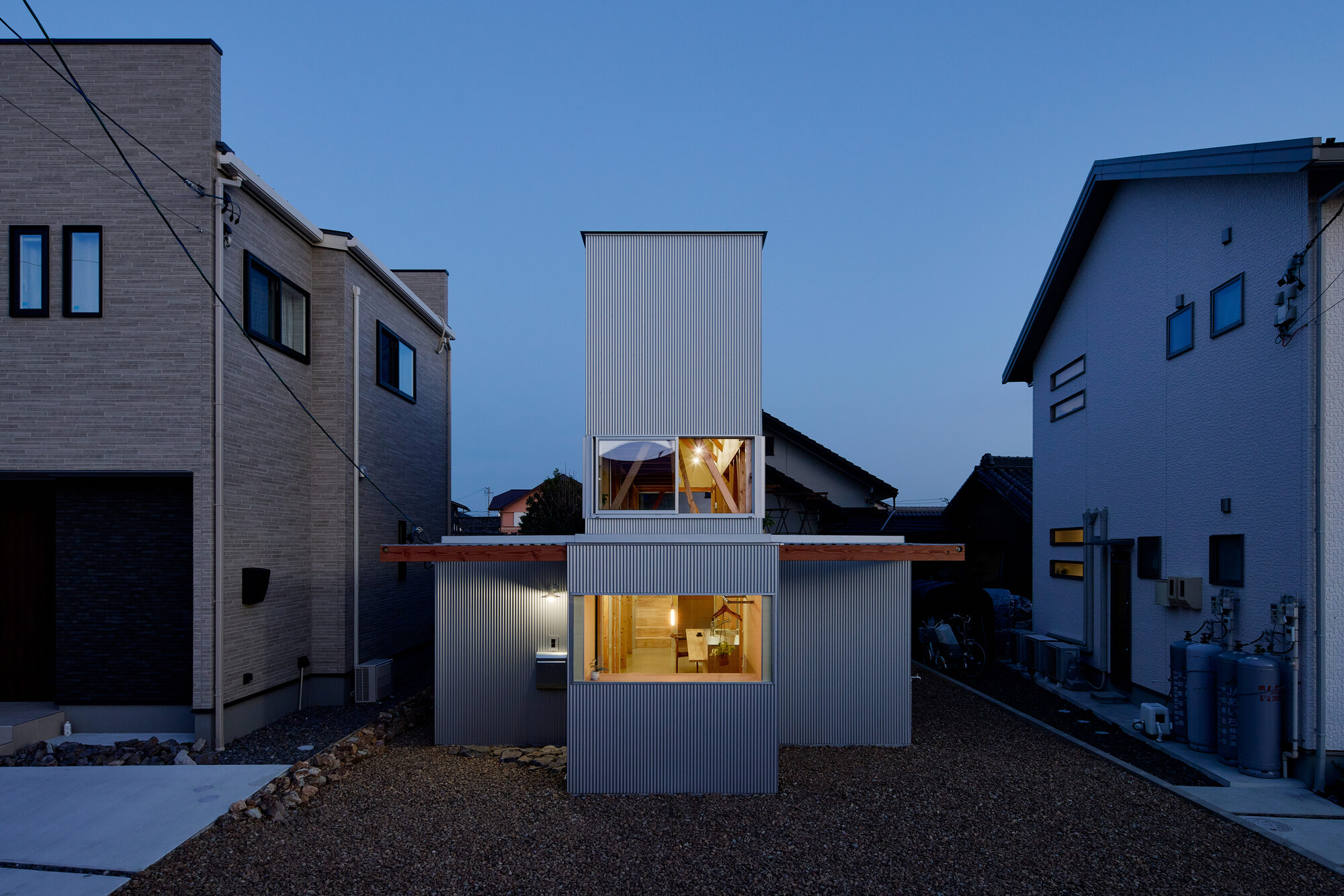Corrugated metal clad volume with illuminated windows at dusk nestled between neighboring houses in gravel yard