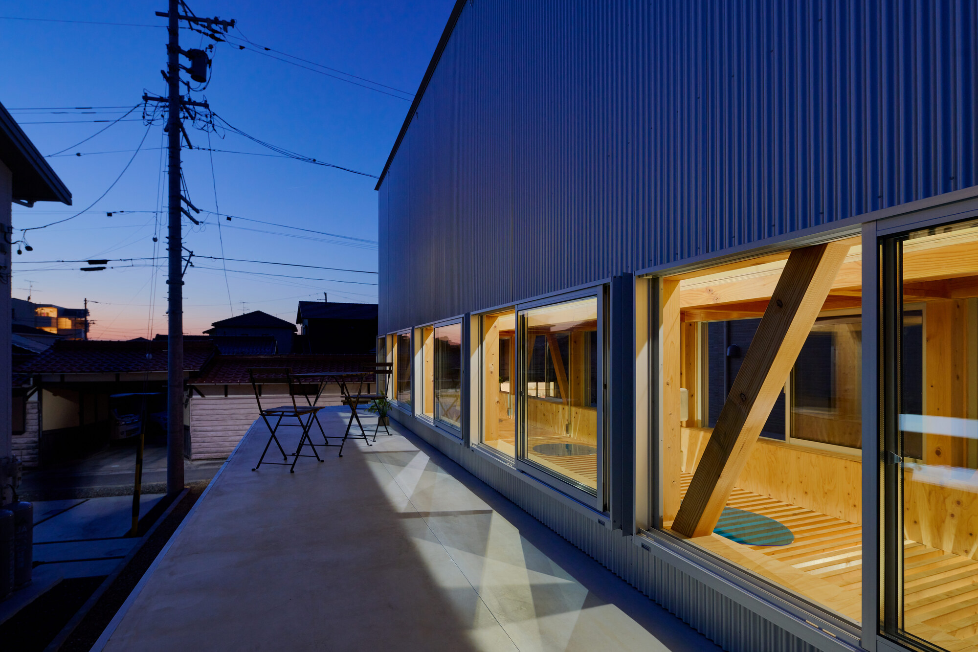 Concrete terrace with folding table beneath corrugated metal facade and glazed timber interior at dusk
