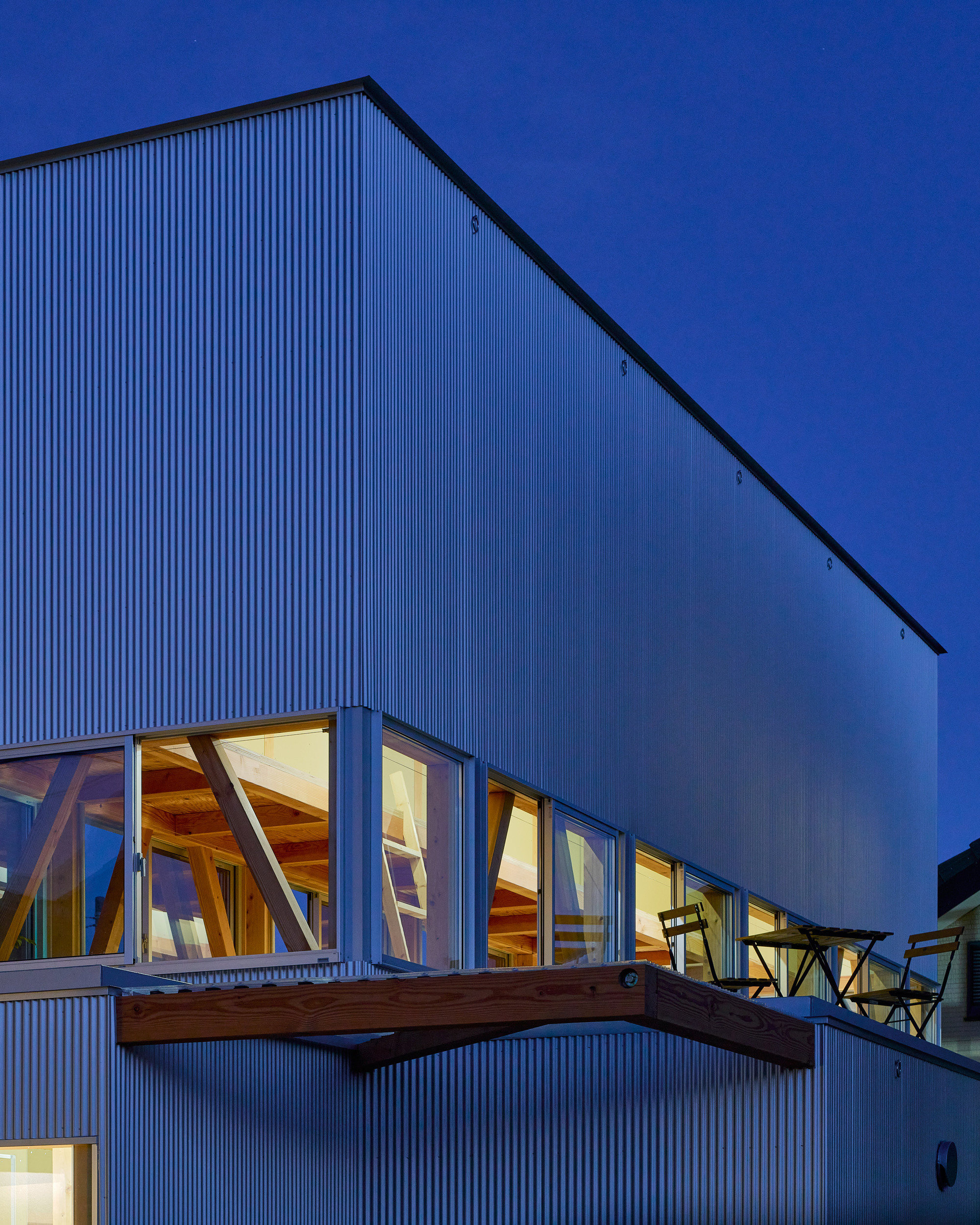 Corrugated metal facade with cantilevered timber deck and angled window openings under blue hour sky