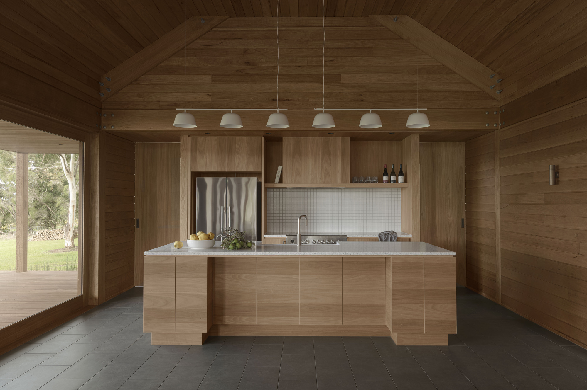 Timber kitchen island with white tile backsplash beneath a vaulted ceiling lined with six pendant lights