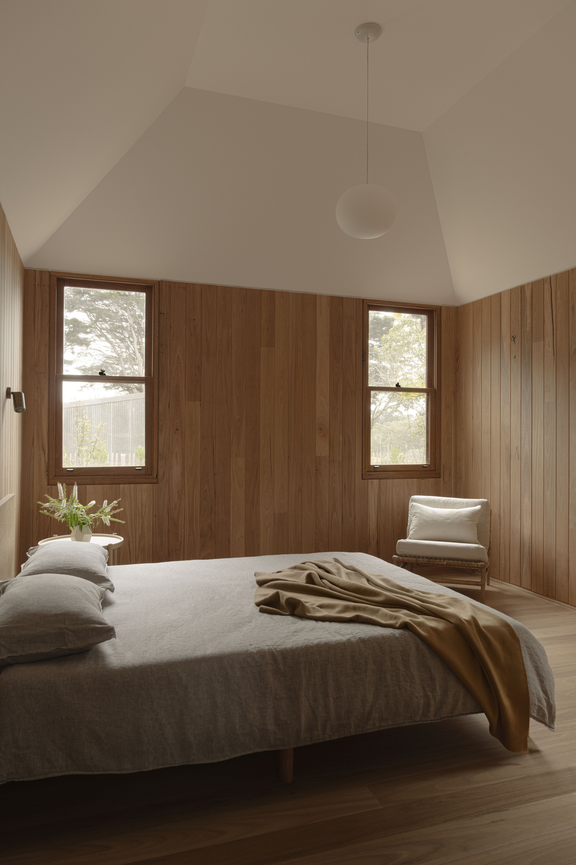 Bedroom with vertical timber paneling, vaulted white ceiling, and two framed windows flanking the bed