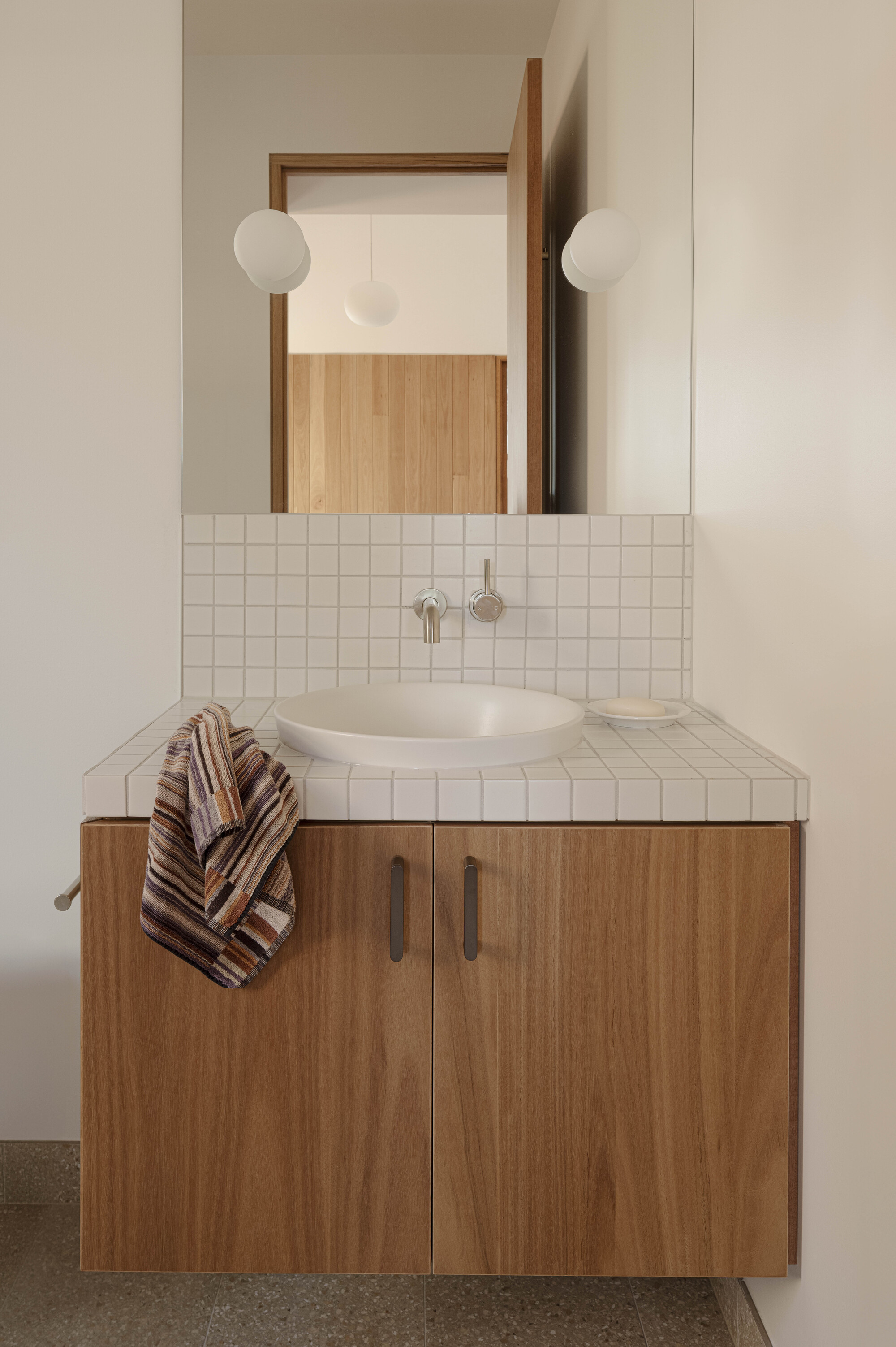 Bathroom vanity with white square tile counter, timber cabinetry and twin globe wall sconces flanking a mirror