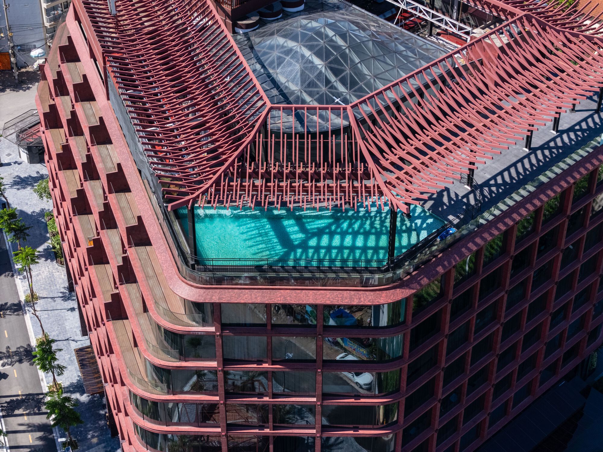 Aerial view of rooftop pool beneath red metal brise-soleil frame and glazed skylight opening