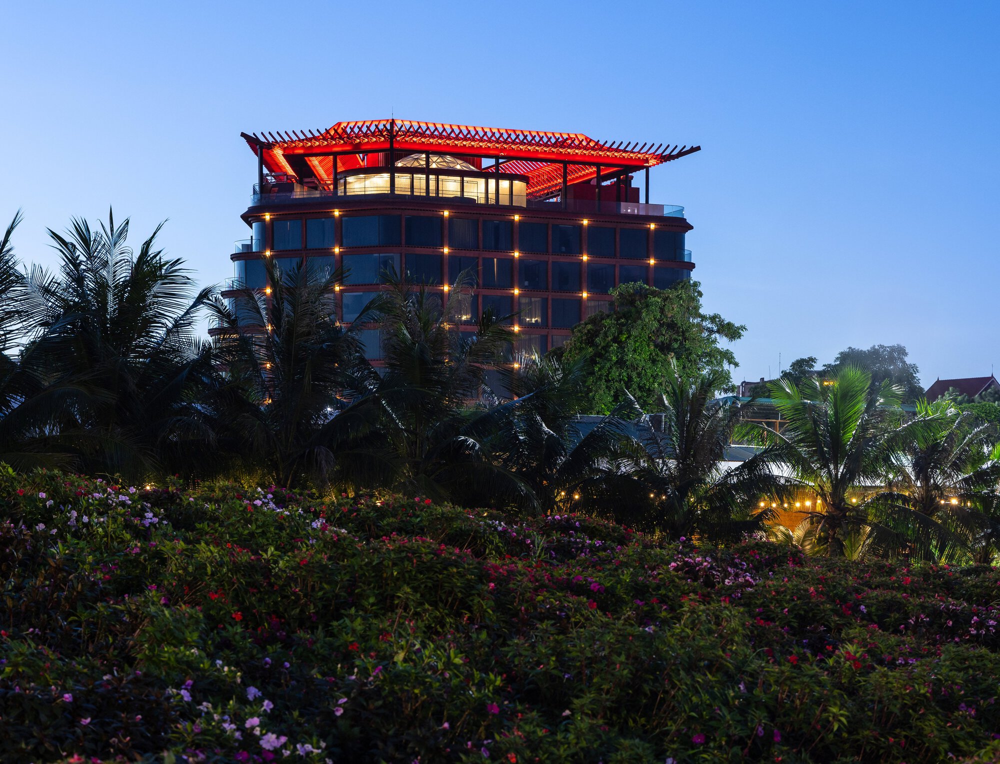 Red lattice roof structure crowning the glazed tower surrounded by palms and flowering plants at evening