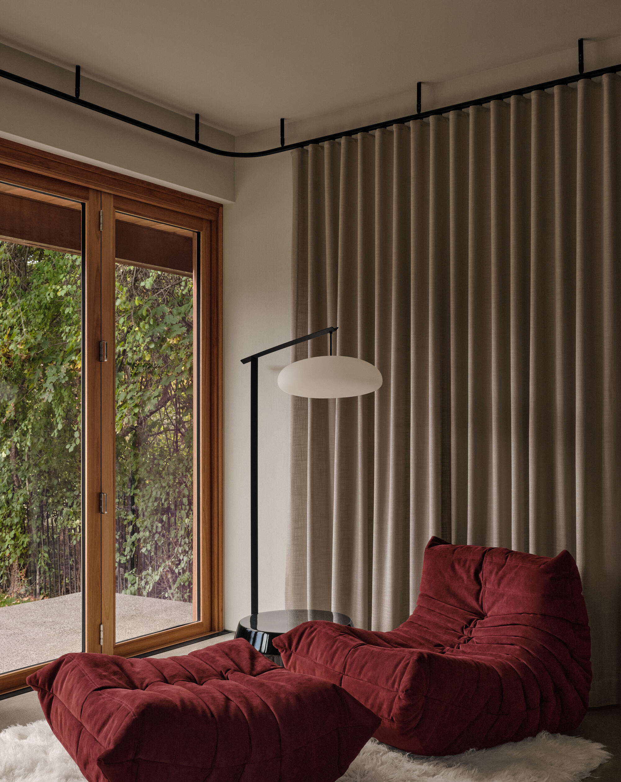 Reading nook with timber-framed glazing, beige curtain track and burgundy upholstered seating overlooking greenery