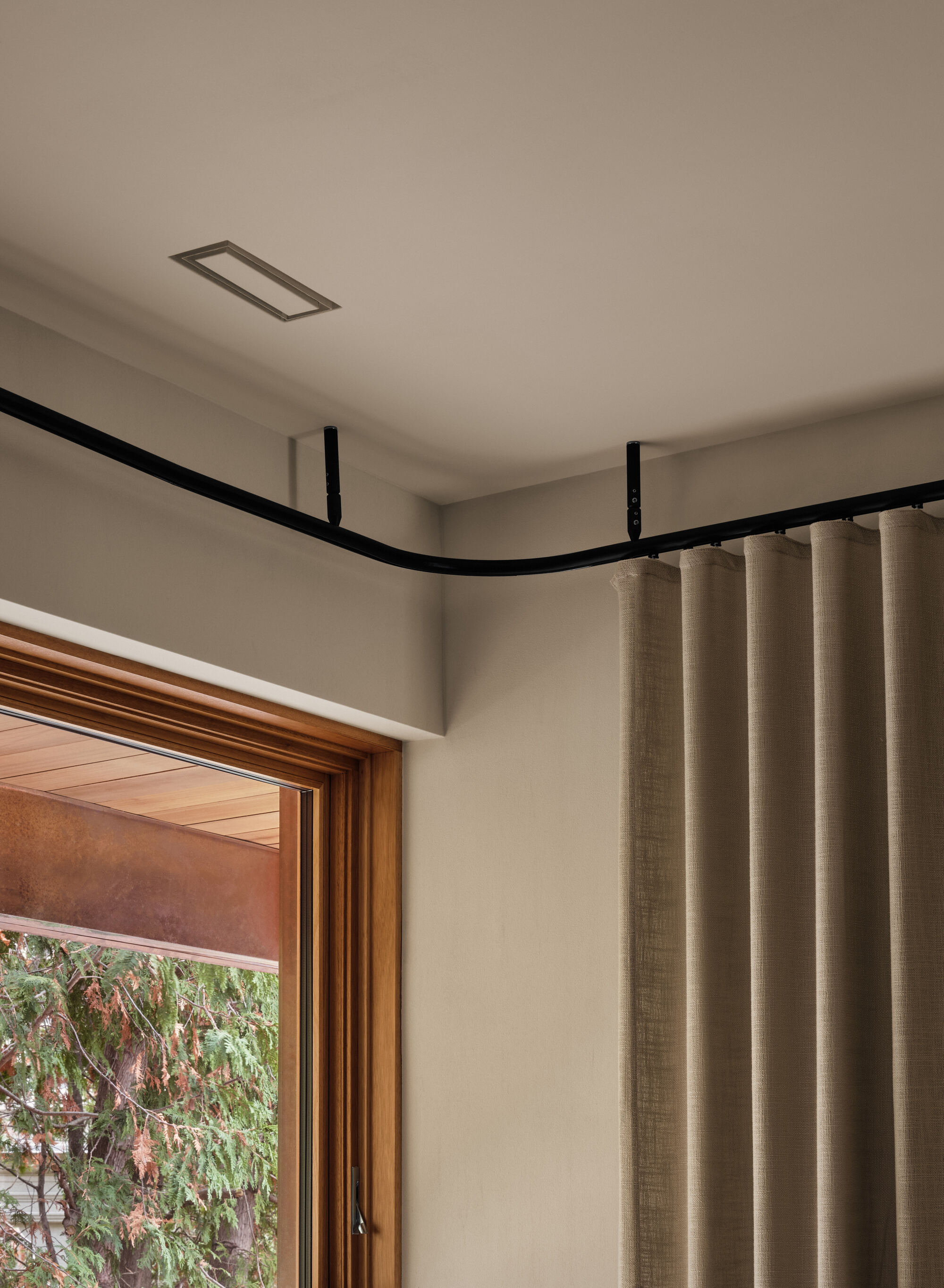 Corner detail of a curved black curtain track above a timber-framed window with garden view