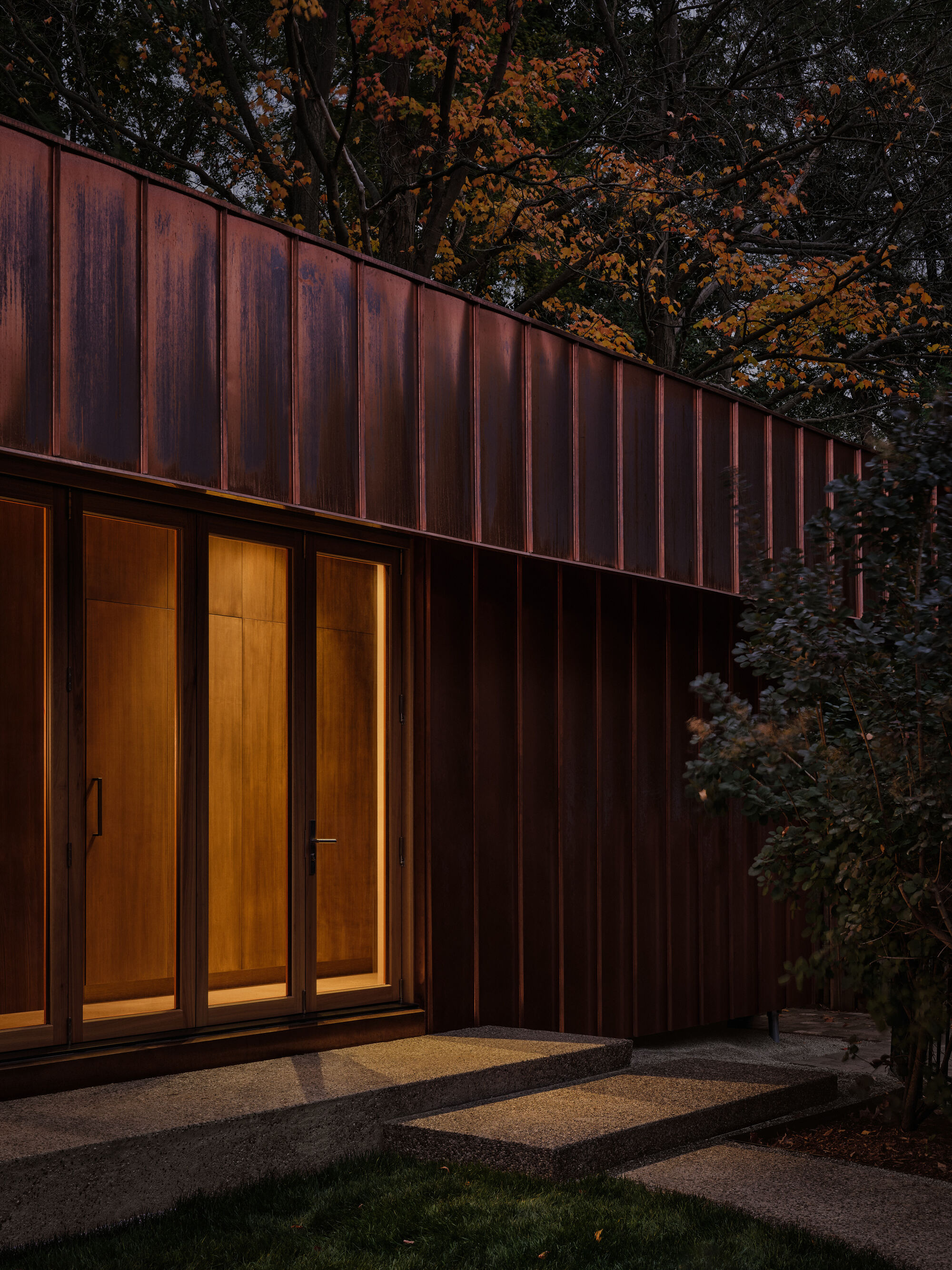 Vertical metal cladding facade with glazed doors illuminated at dusk beneath autumn trees