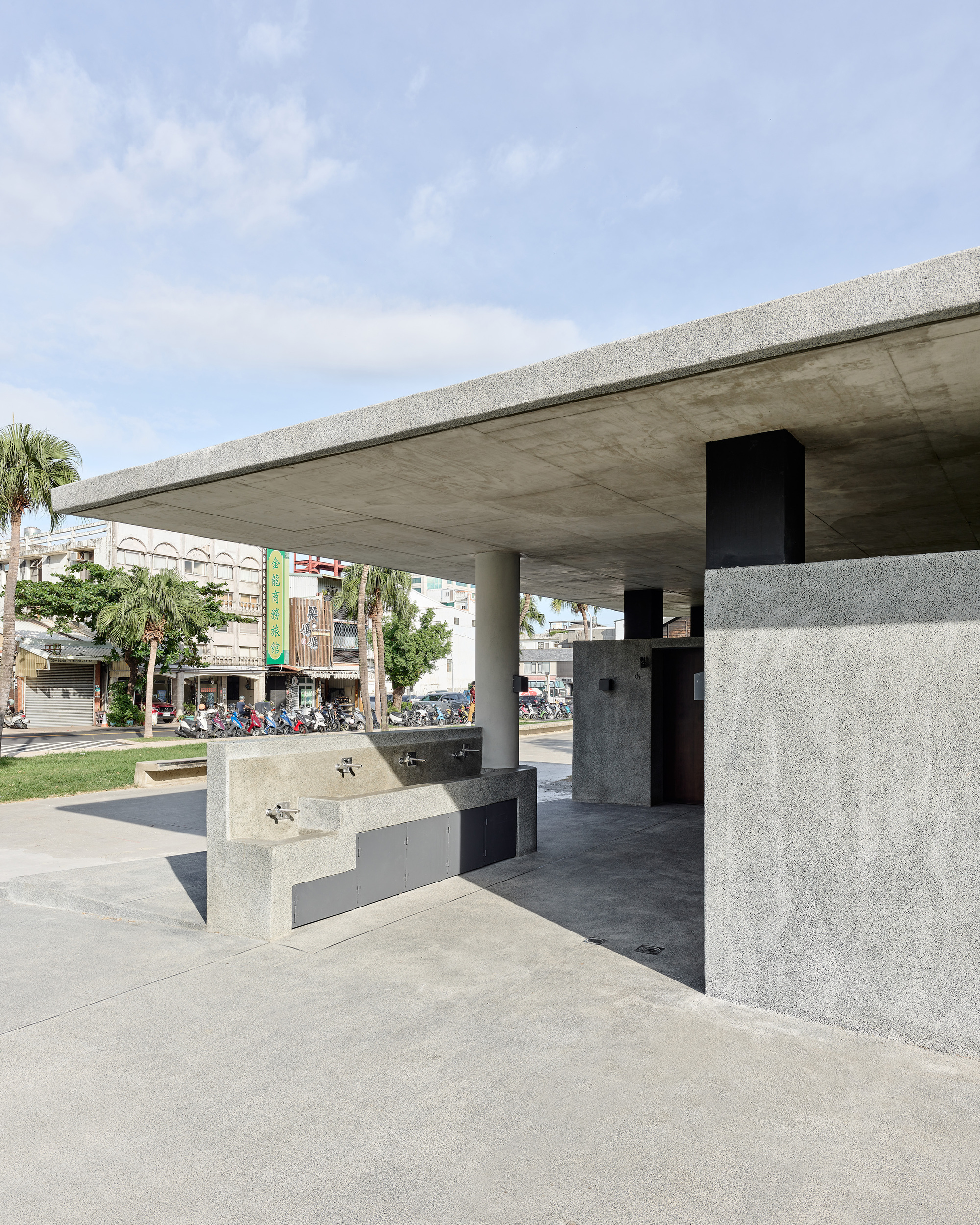 Covered pavilion with concrete columns and exposed soffit panels overlooking a public plaza with street beyond